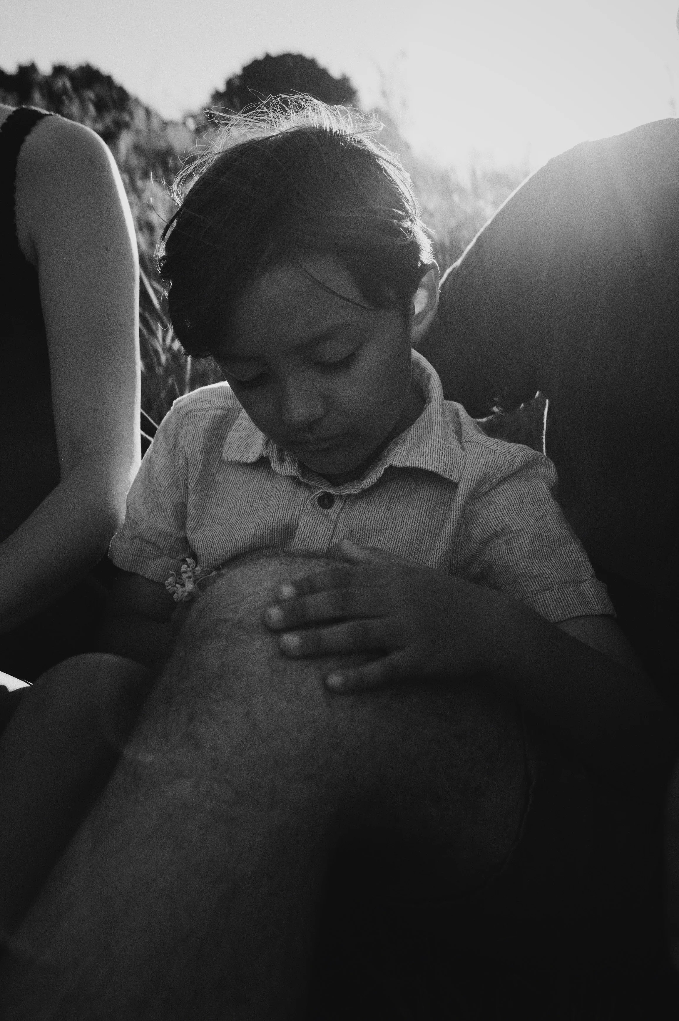 A black-and-white photo of a young boy gently holding a person's leg in an outdoor setting during sunset, with trees in the background.