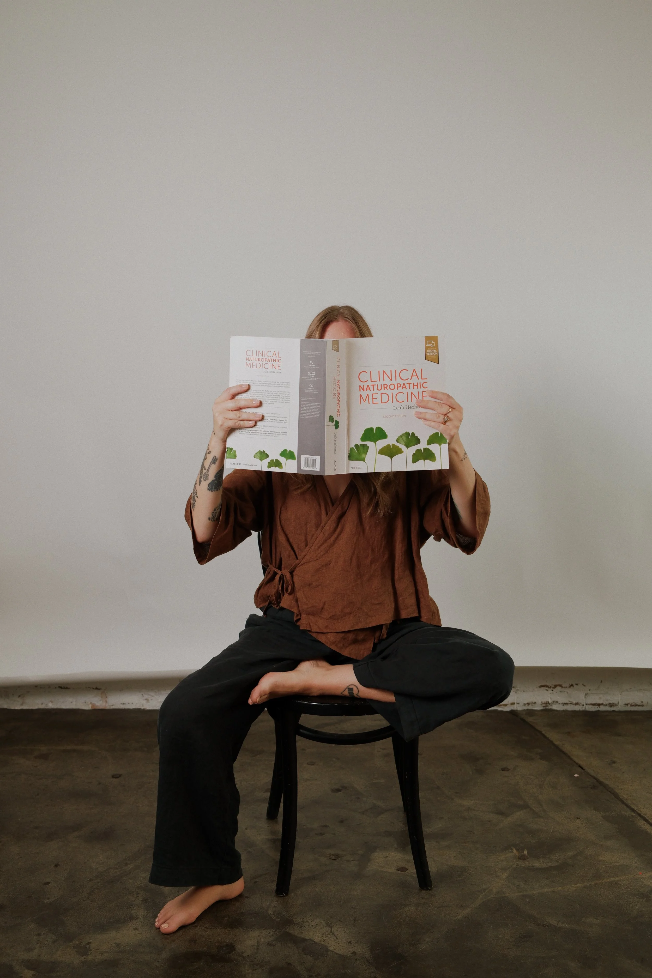 Person sitting cross-legged on a chair, barefoot, wearing a brown top and black pants, holding a book titled 'Clinical Naturopathic Medicine' in front of their face, against a plain wall.