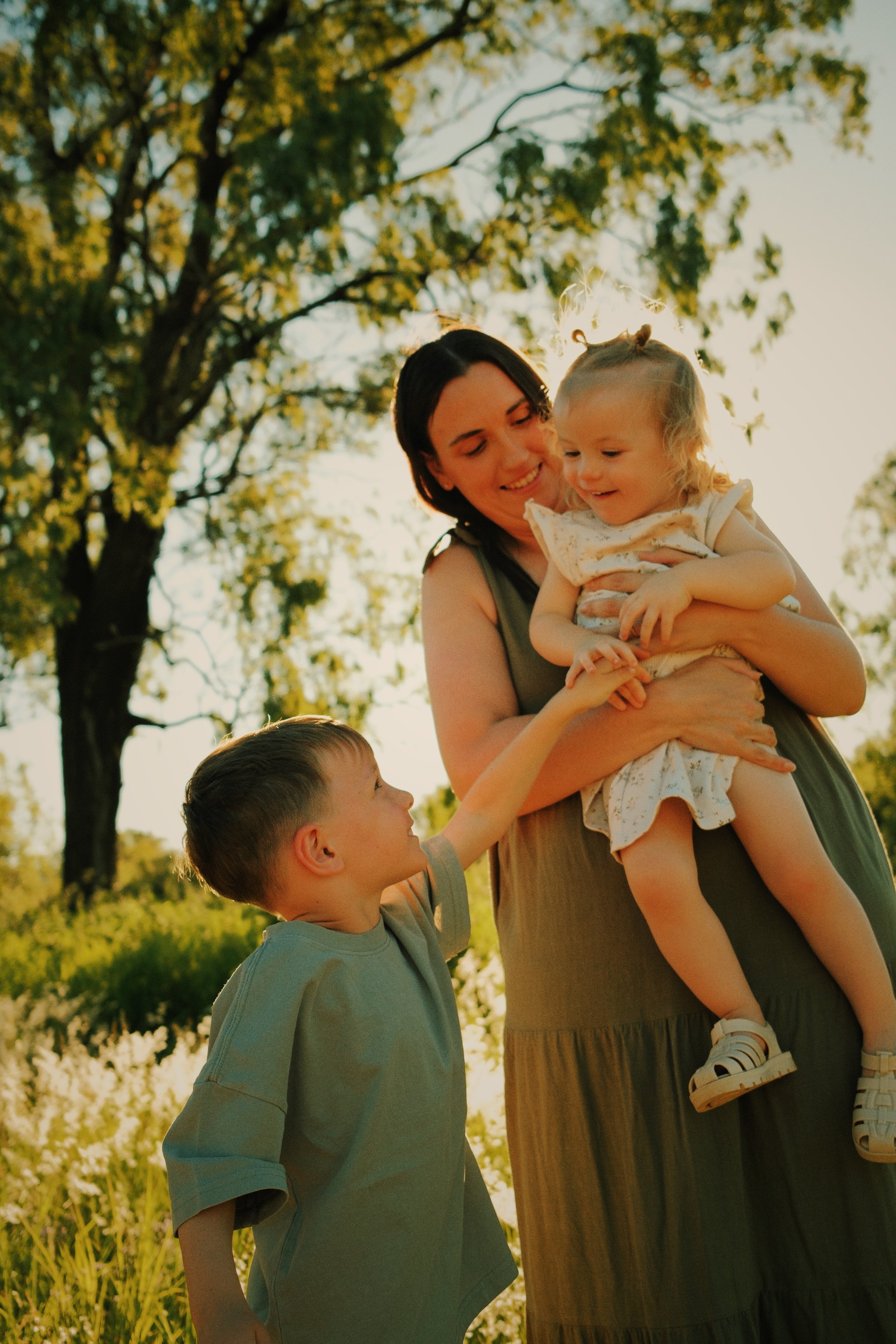 A woman holding a young girl in a field with two children playing and trees in the background during sunset.
