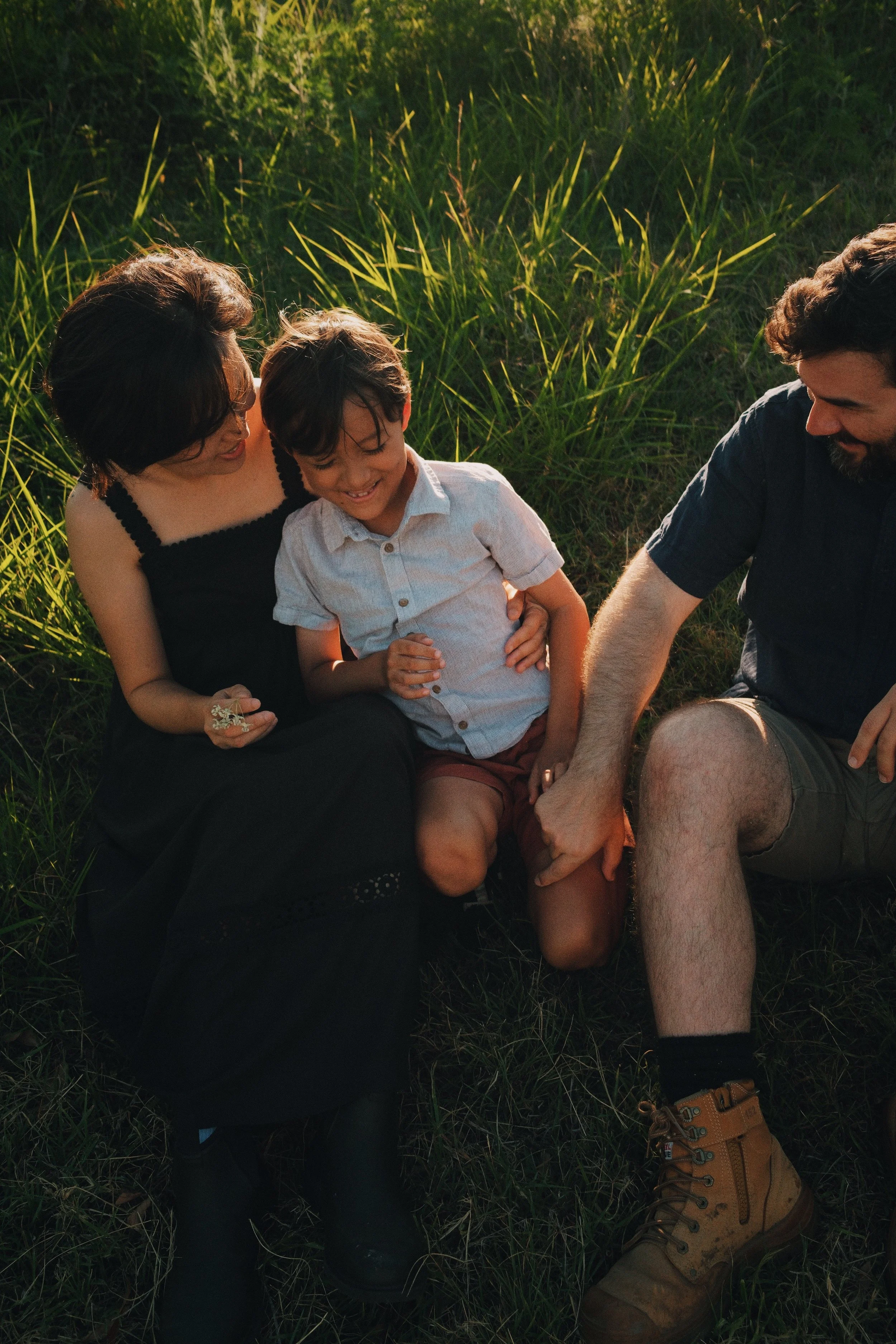 A family sitting on the grass in a field, with the mother, father, and young boy smiling and holding hands.
