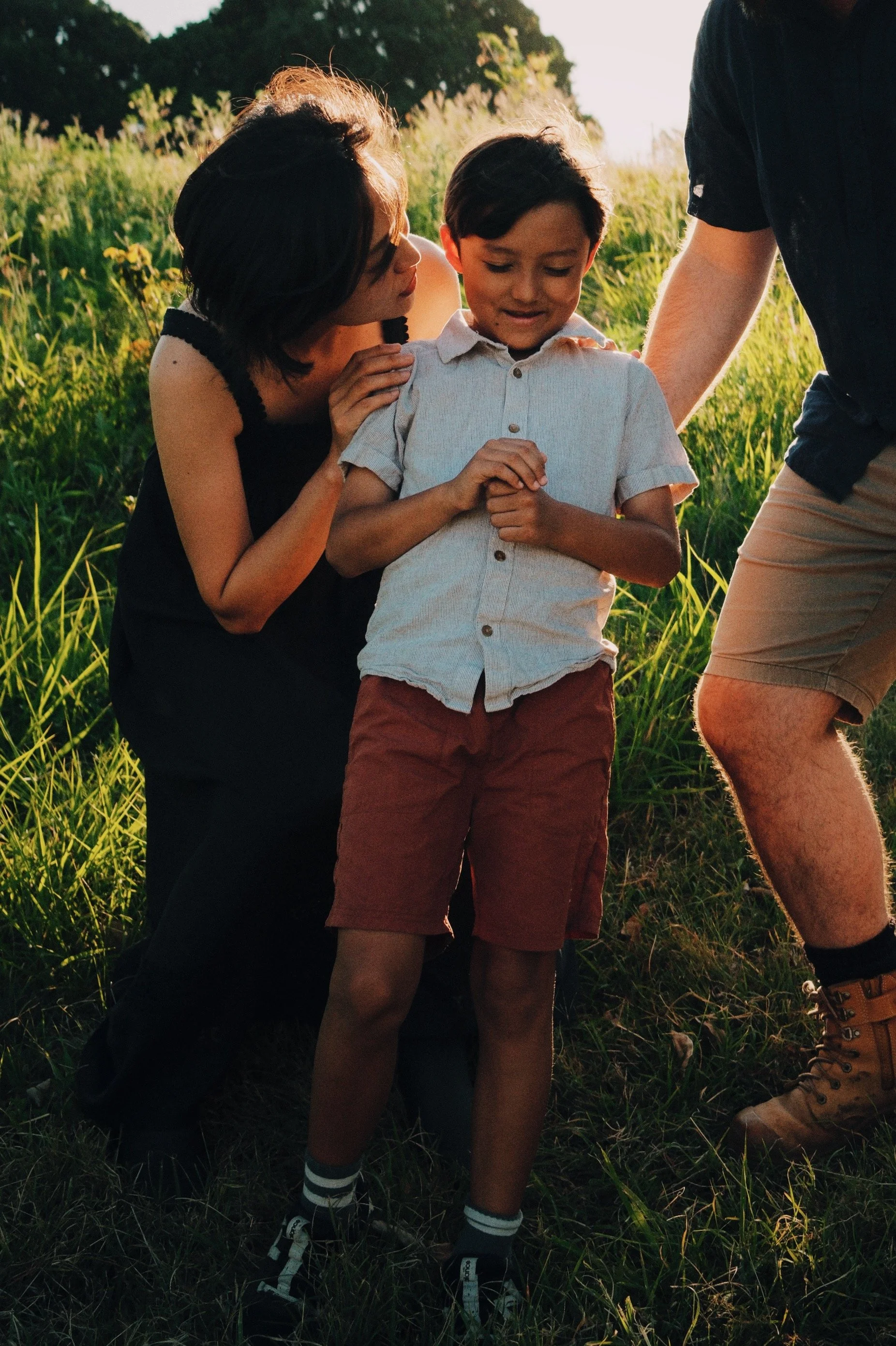 A woman and two men helping a young boy walk through tall grass outdoors in the late afternoon sunlight.