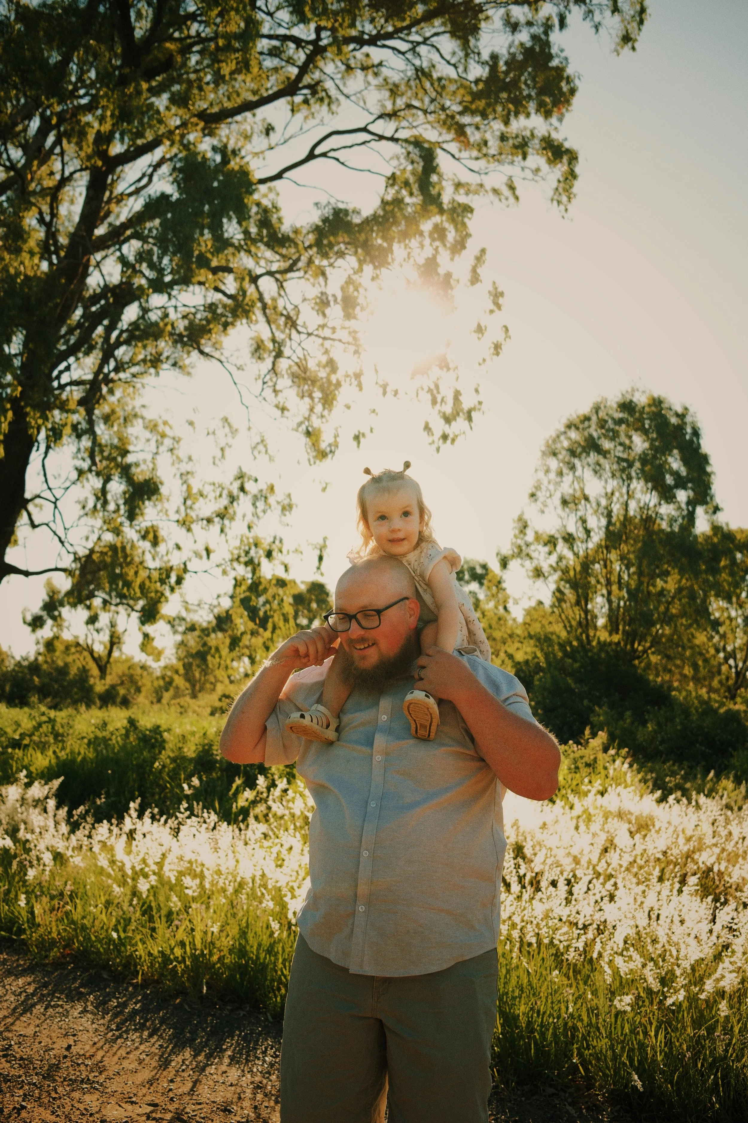 A man with glasses and a beard carries a young girl on his shoulders in a sunlit outdoor setting with tall grass and trees.