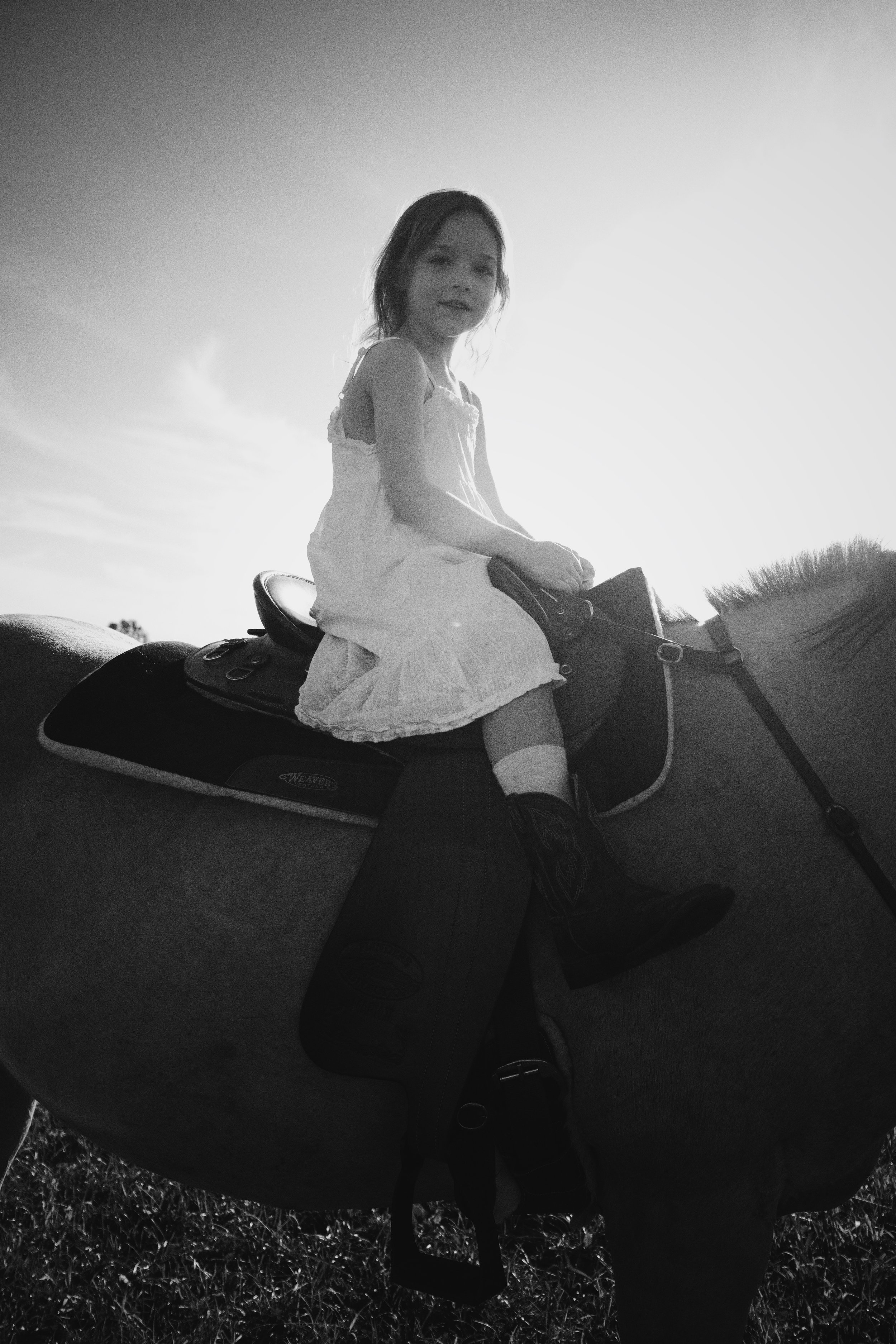 A young girl in a white dress sitting on a horse with a saddle, outdoors during sunset.