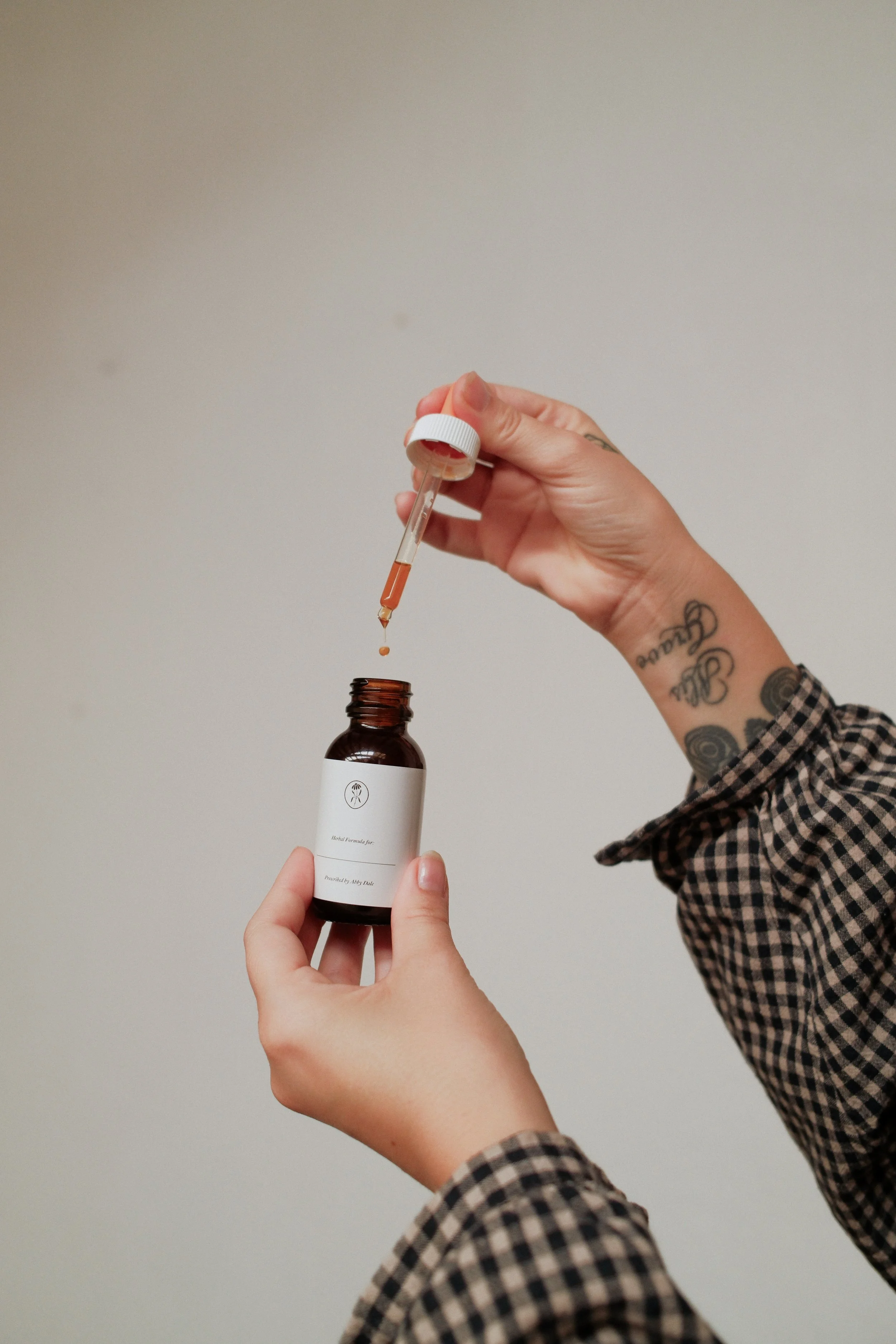 Person holding a small brown bottle with a white label and a dropper, dispensing a brown liquid into the bottle.