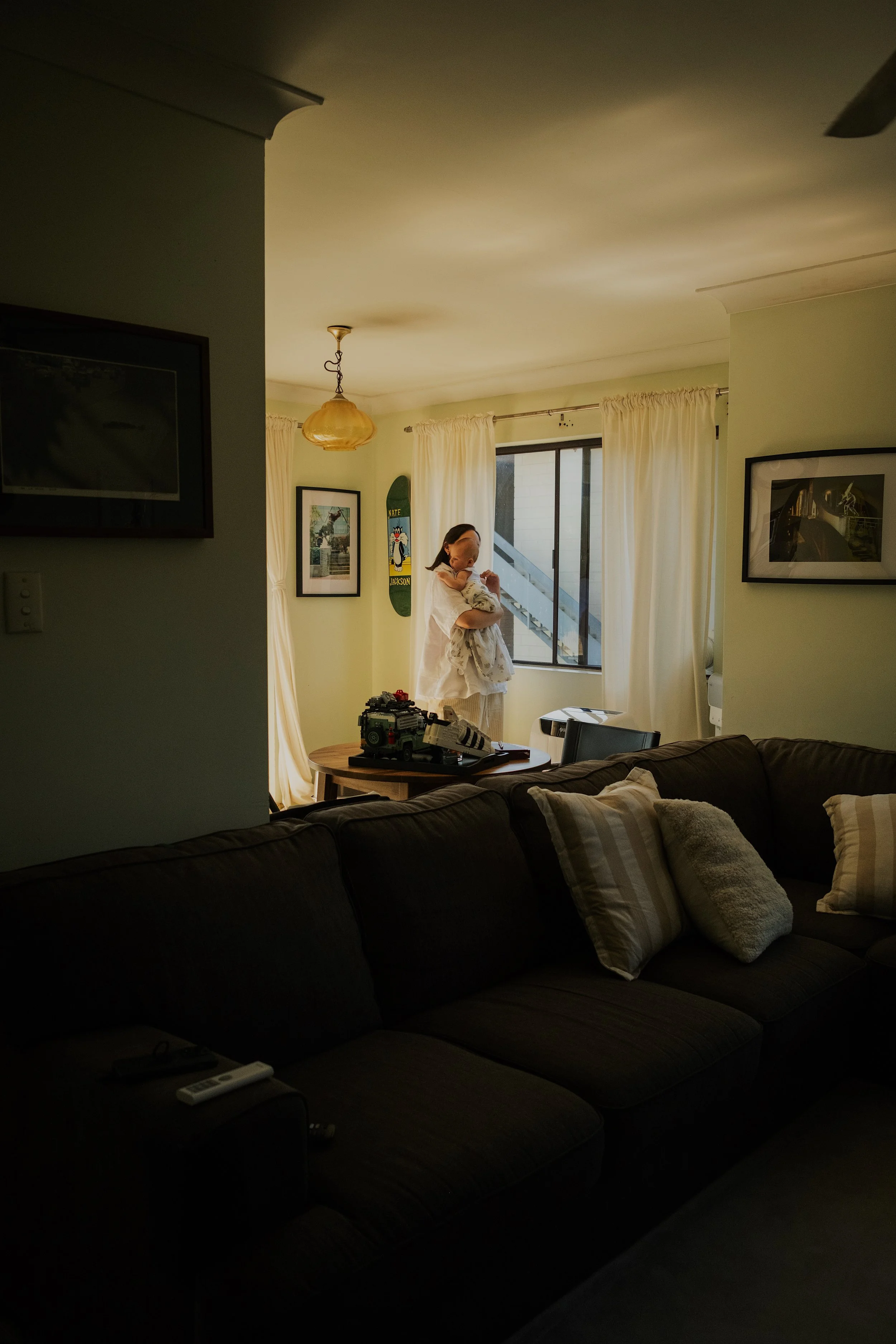 A woman holding a small dog standing near a window in a living room with white curtains, framed pictures on the wall, and a skateboard leaning against the wall.