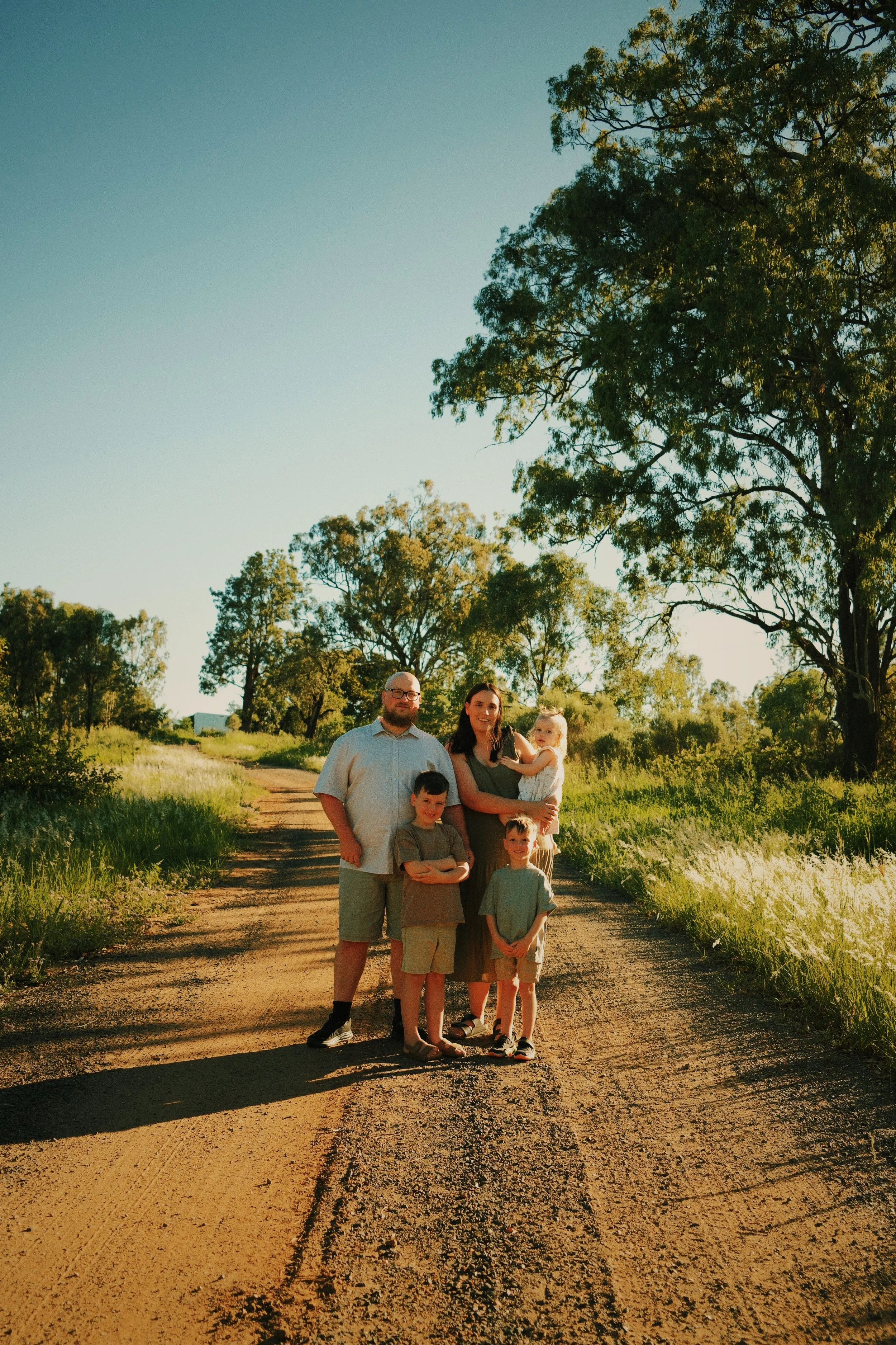 A family of five stands on a dirt path in a rural area with tall grass and trees, under a clear blue sky, during sunset.