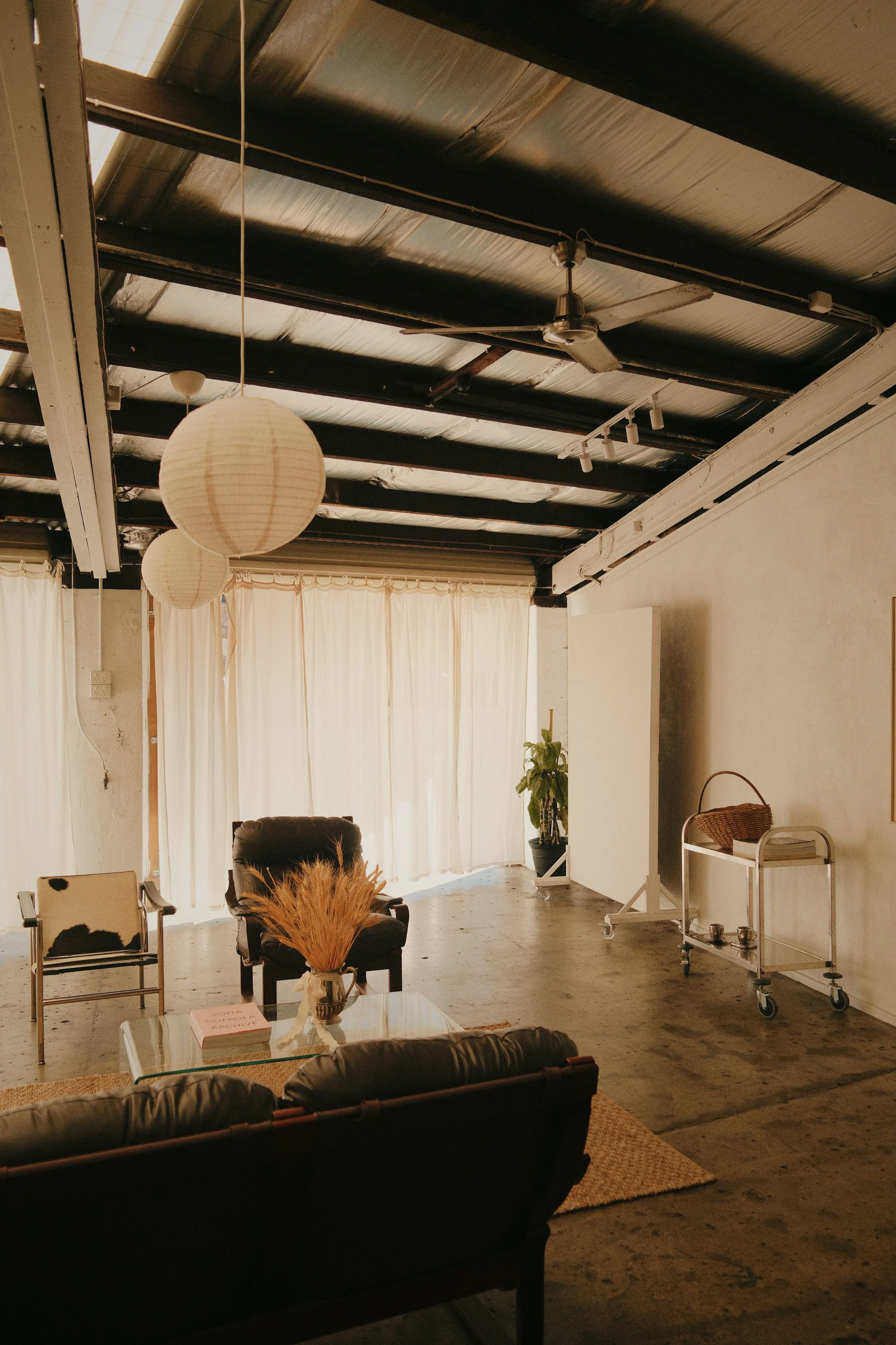 Interior of a room with industrial ceiling, beige curtains, and simple furniture including chairs, a glass table with a vase of dried plants, and a wheelchair cart.