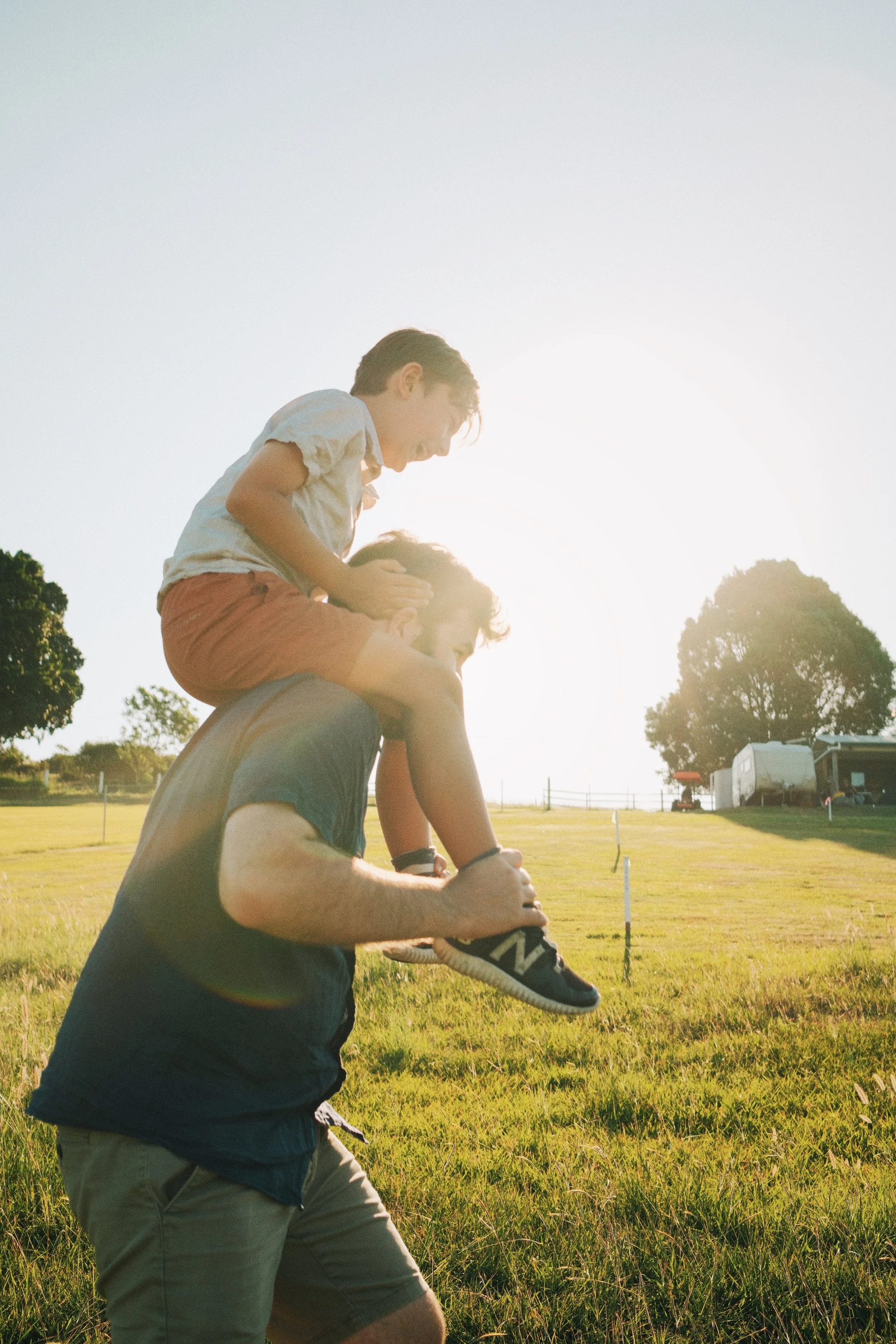 A man is carrying a young boy on his shoulders outdoors in a grassy field during sunset.