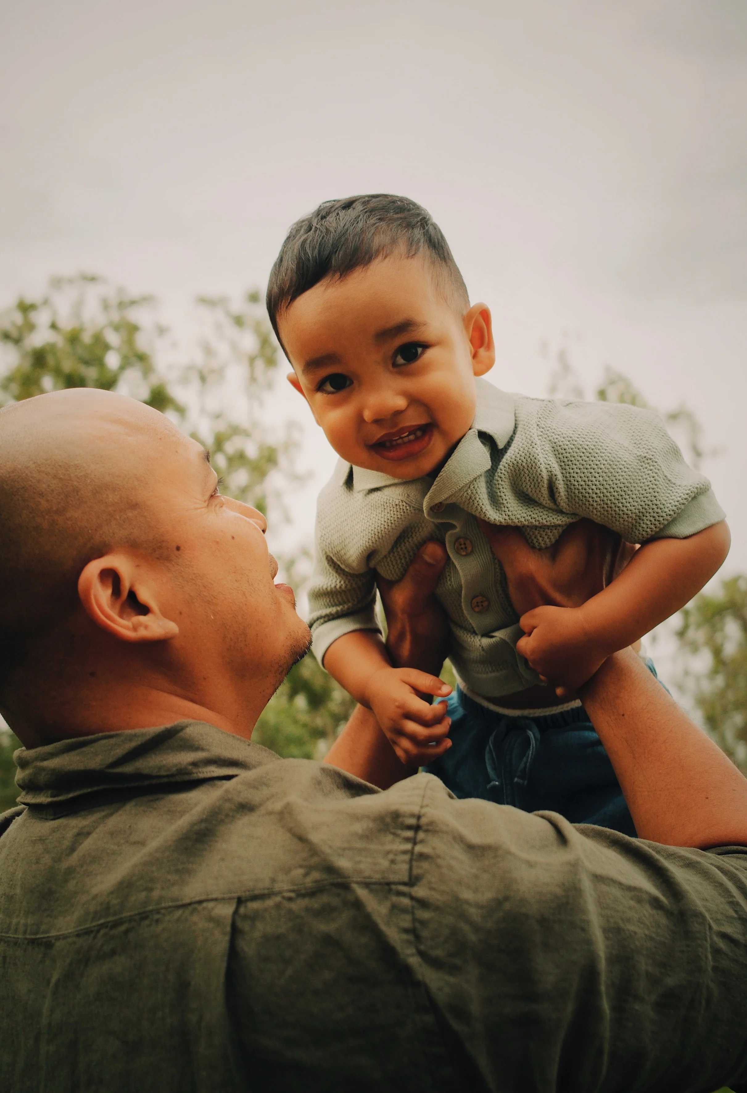 A man holding a young boy up in the air outdoors, with trees and a cloudy sky in the background.