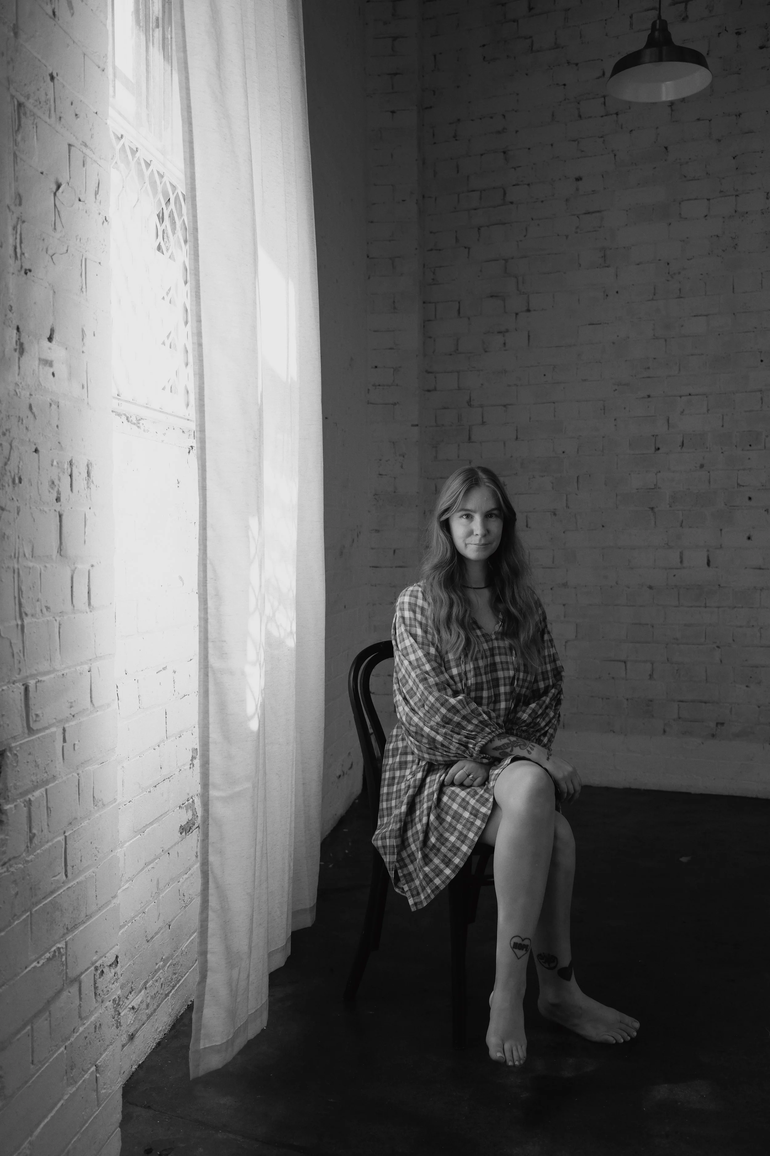 A young woman with long wavy hair sitting barefoot on a chair in front of a brick wall. She is wearing a plaid dress and has tattoos on her legs. Natural light coming through a window illuminates the scene.