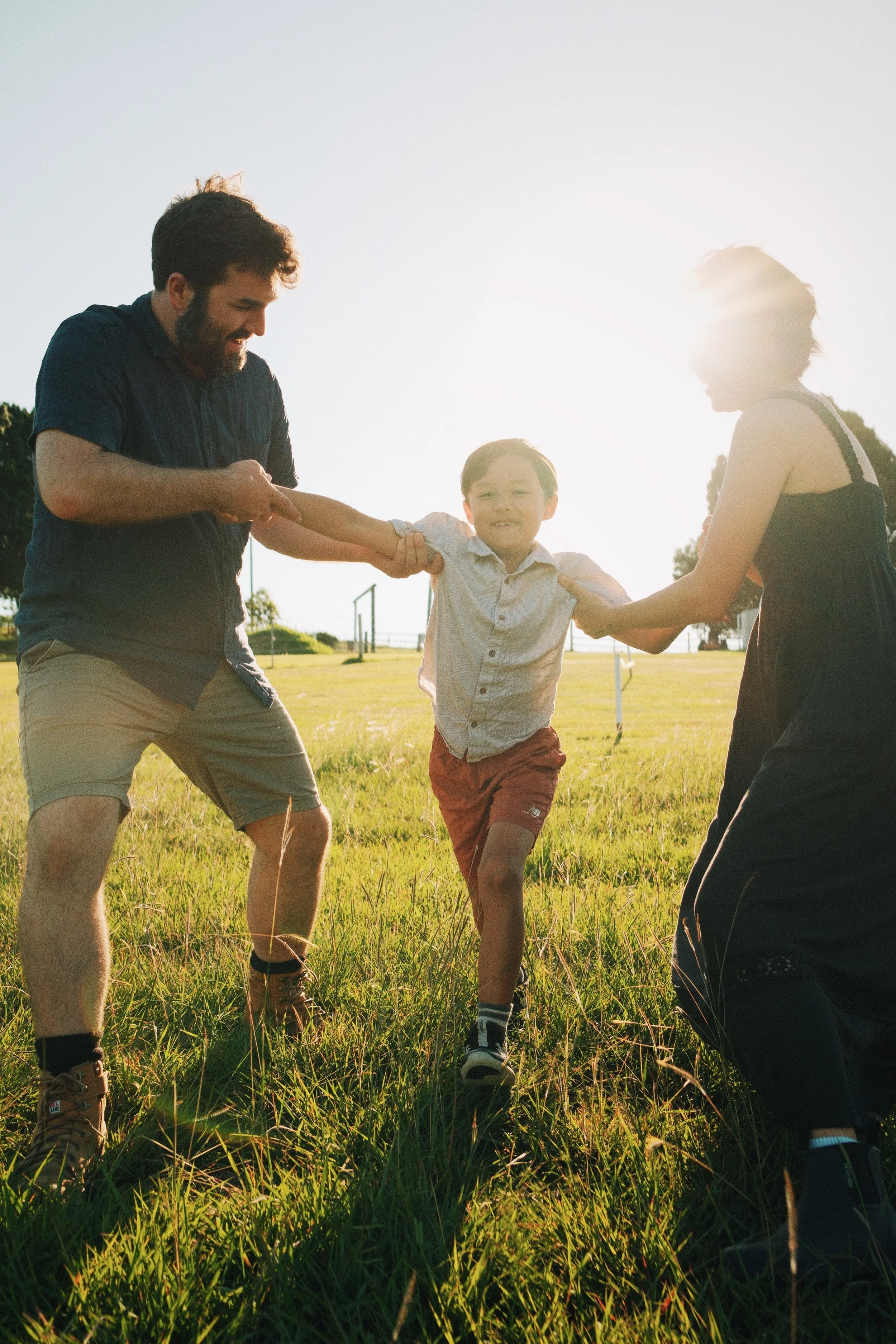 A boy is being playfully pulled in opposite directions by two adults in a grassy field during sunset.