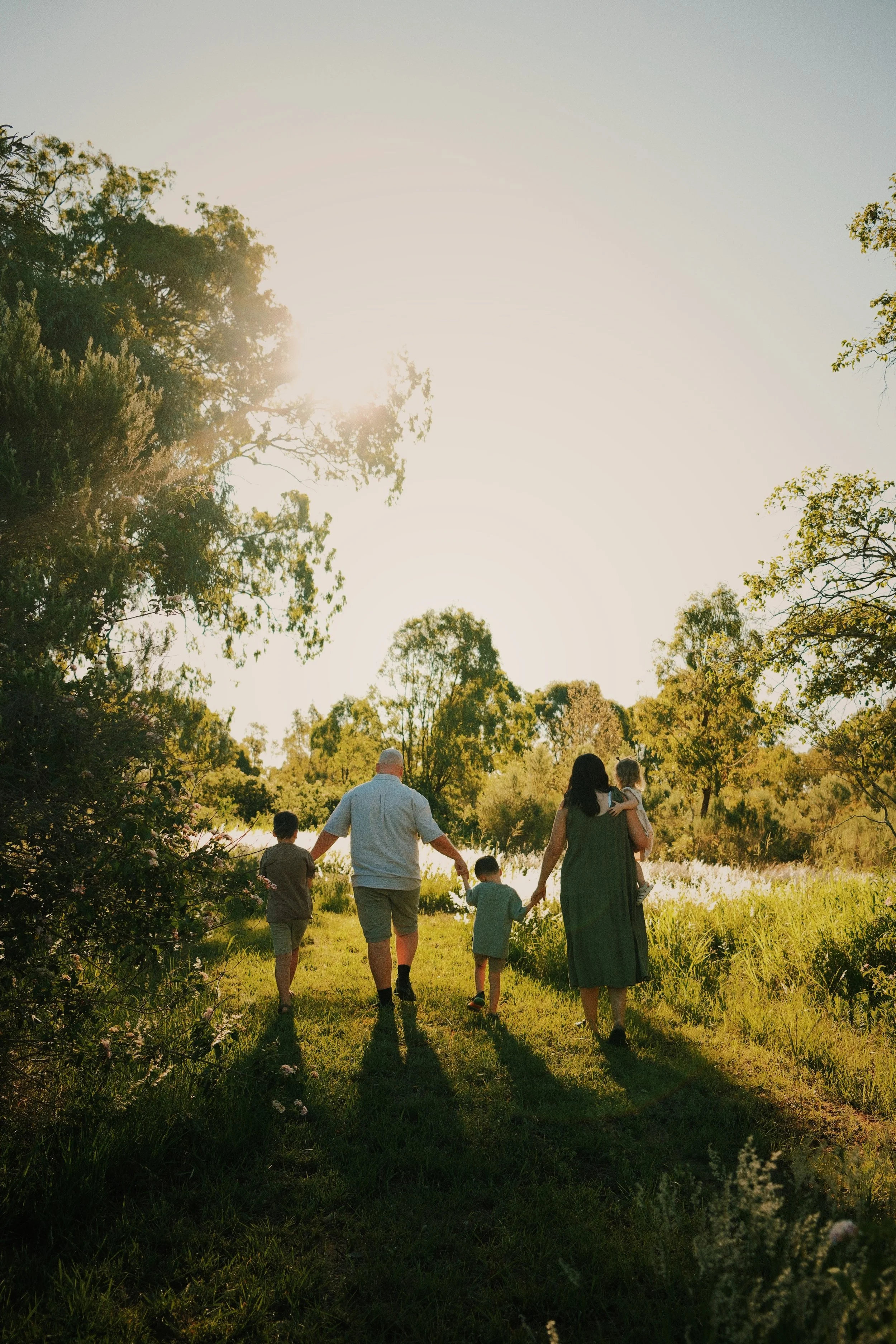 A family of five walking hand in hand on a grassy trail through a wooded area during sunset.