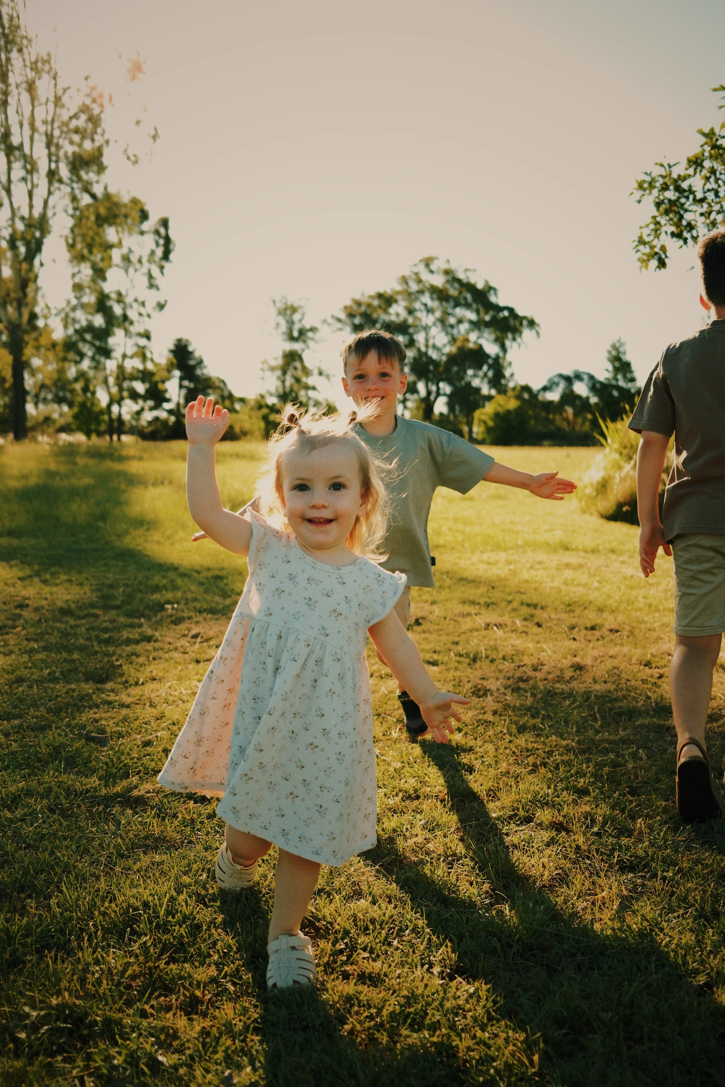 Three children, a girl and two boys, running in a grassy park on a sunny day with trees in the background, smiling and enjoying themselves.