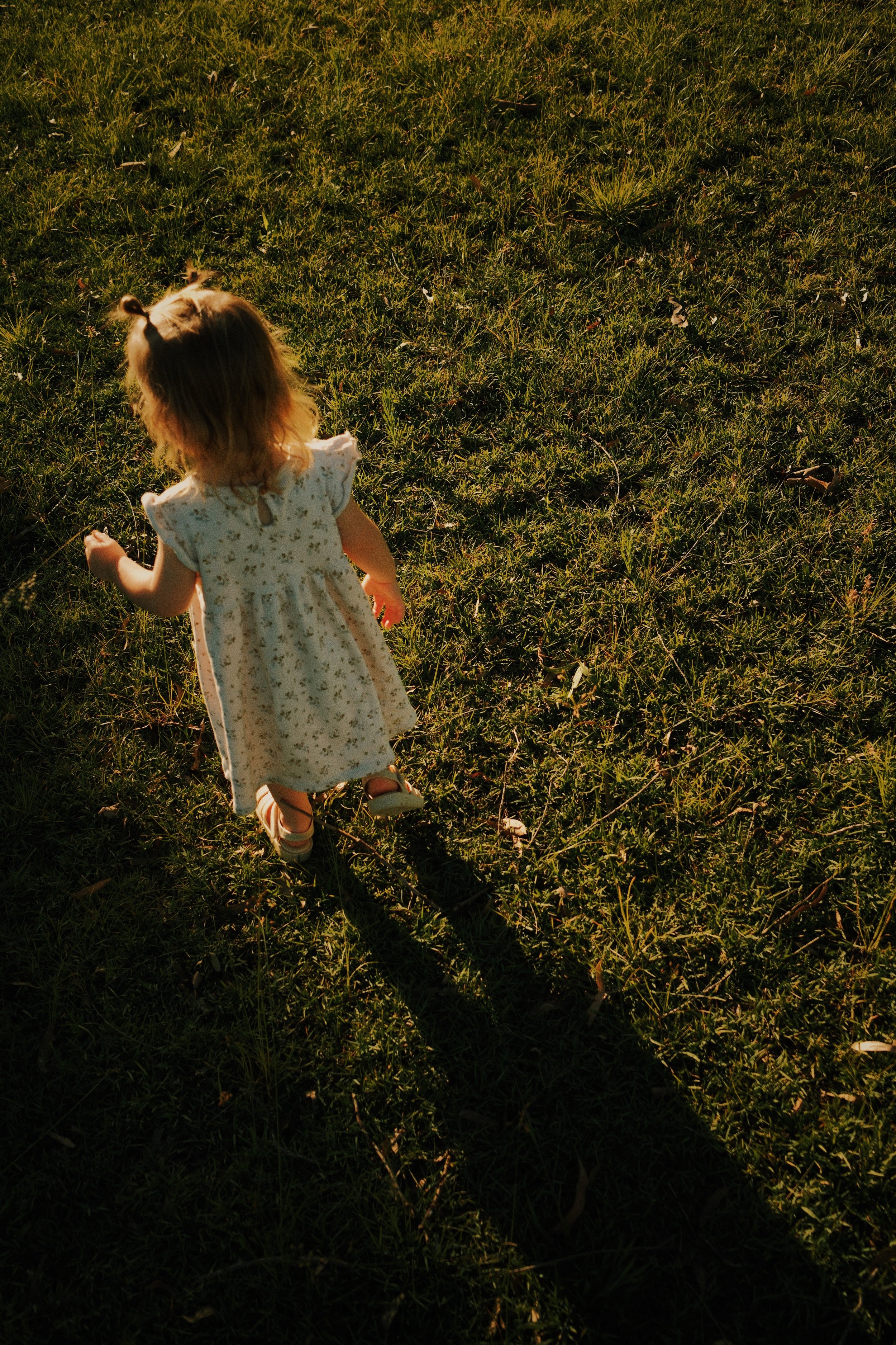 A young girl with blonde hair in pigtails, wearing a light-colored floral dress and shoes, walking on grass during sunset, casting a long shadow.