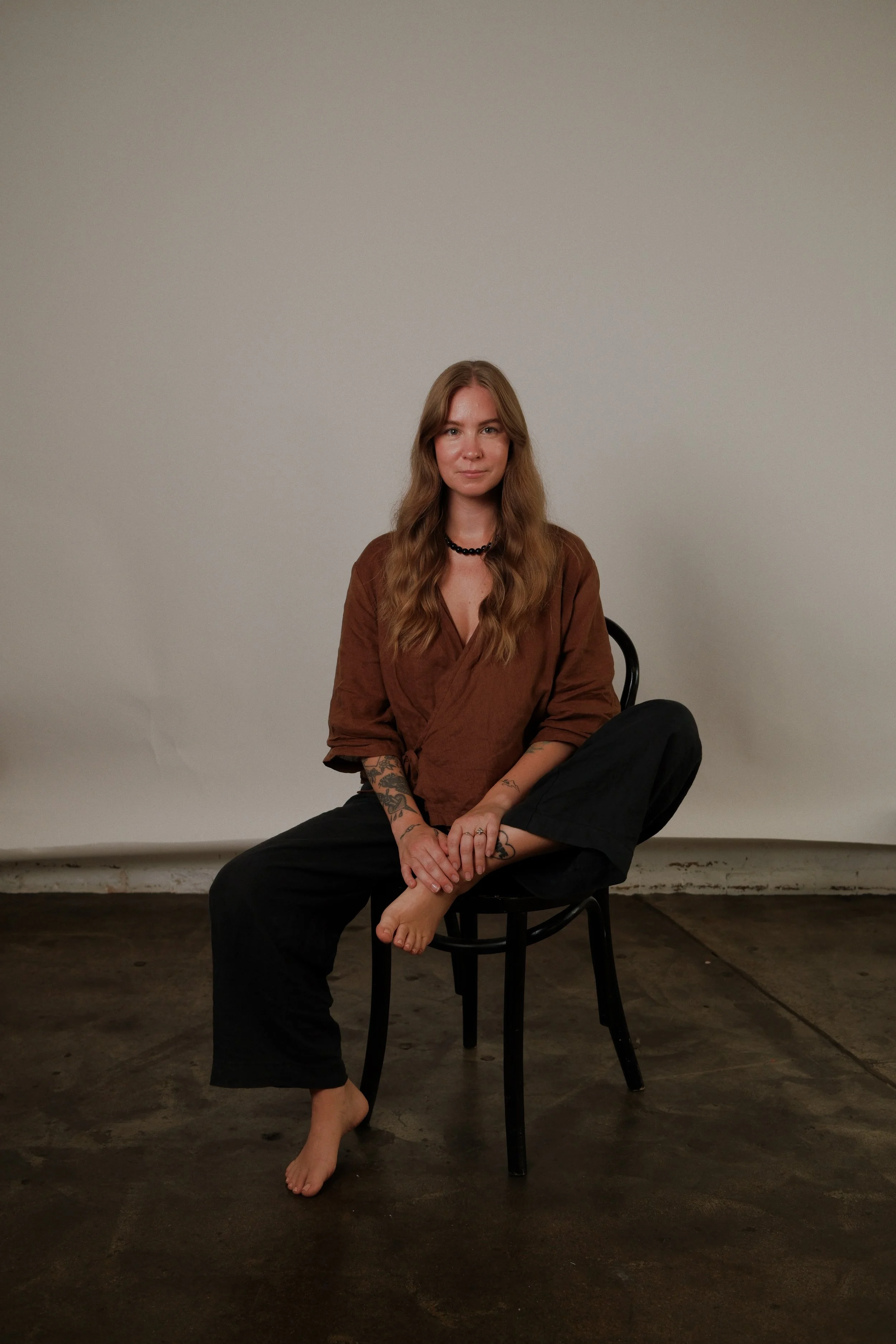 A woman with long wavy hair, tattoos on her arms, wearing a brown top and black pants, sitting barefoot on a black chair against a plain light background.