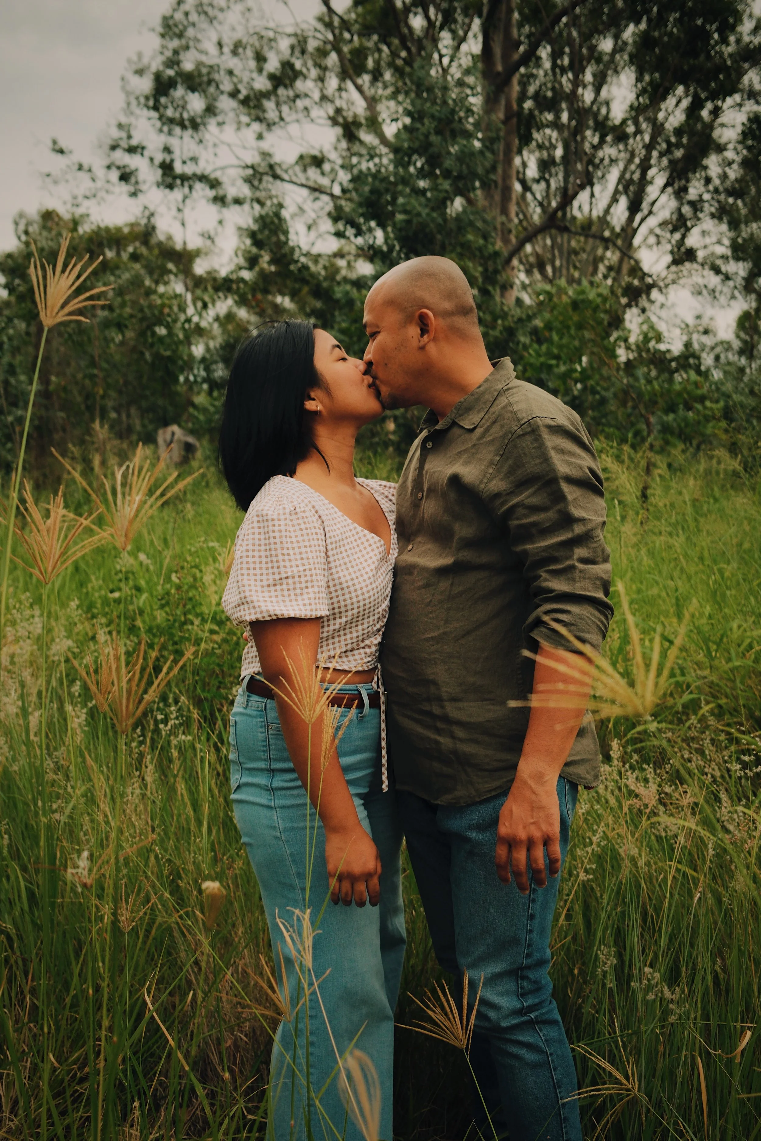 A couple kissing in a field of tall grass with trees in the background.