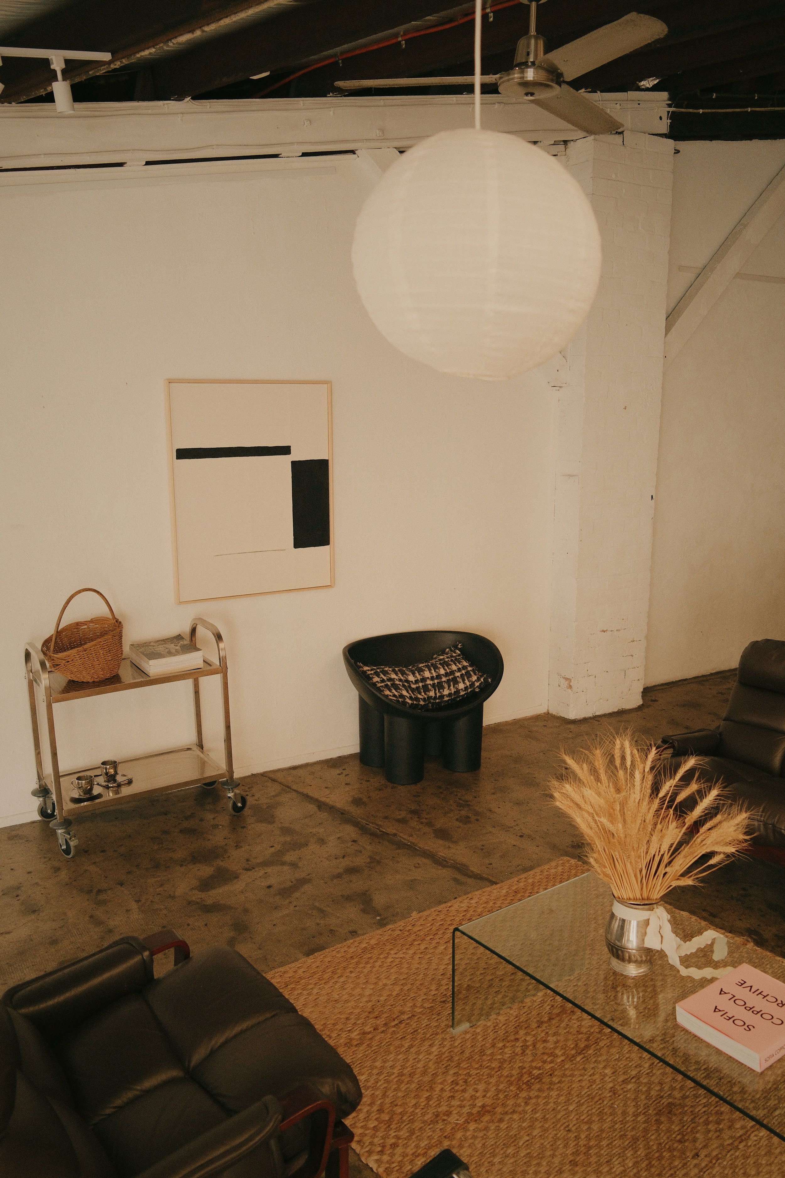 Modern living room featuring a white wall with minimalist black and white artwork, a black chair with a patterned cushion, and a cart with a wicker basket and magazines. A glass coffee table with a vase of dried wheat and a book is in front of a leat