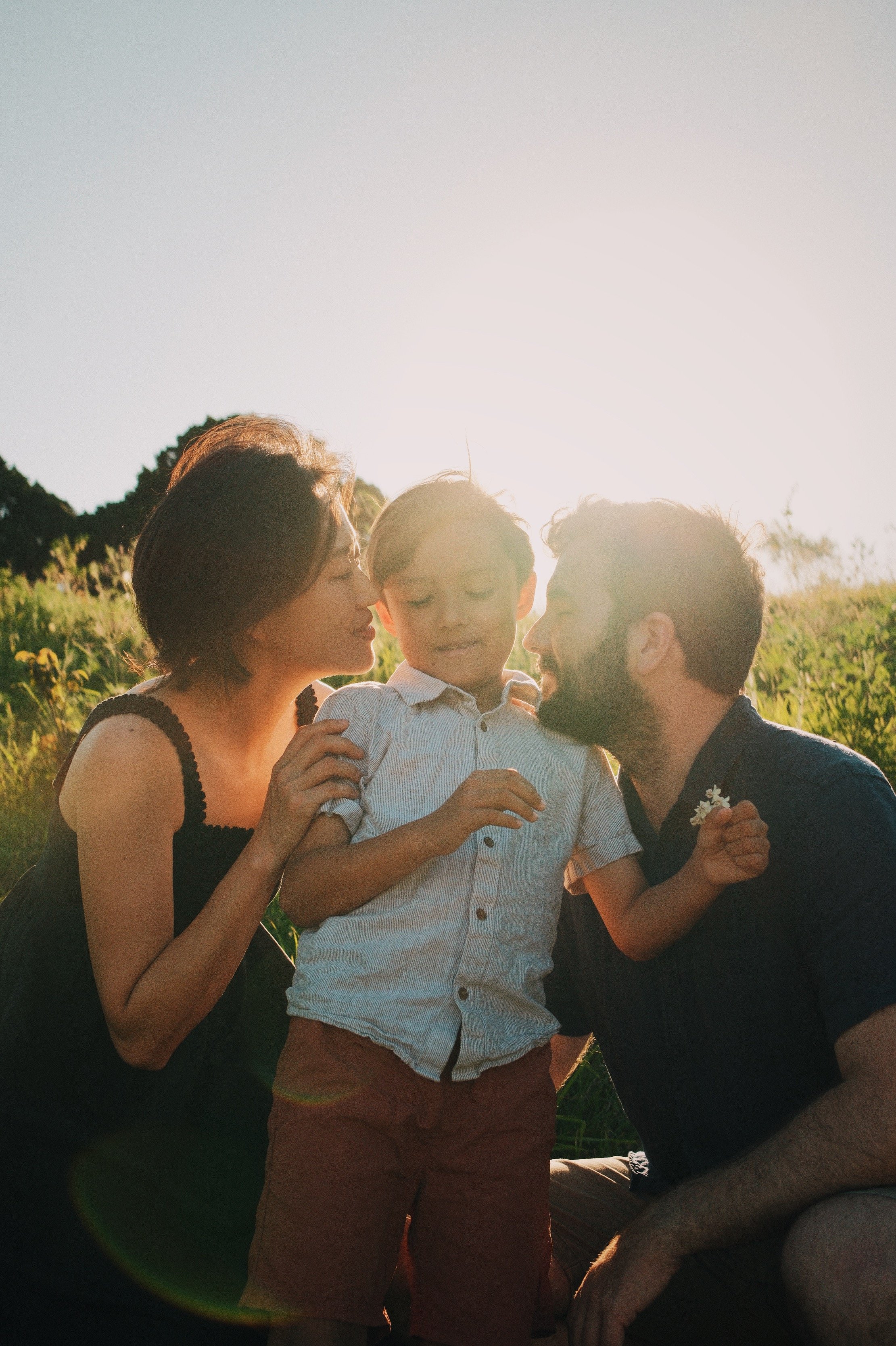 A family of three, a woman, a man, and a young boy, sharing a moment outdoors during sunset, with the sun behind them, surrounded by greenery.