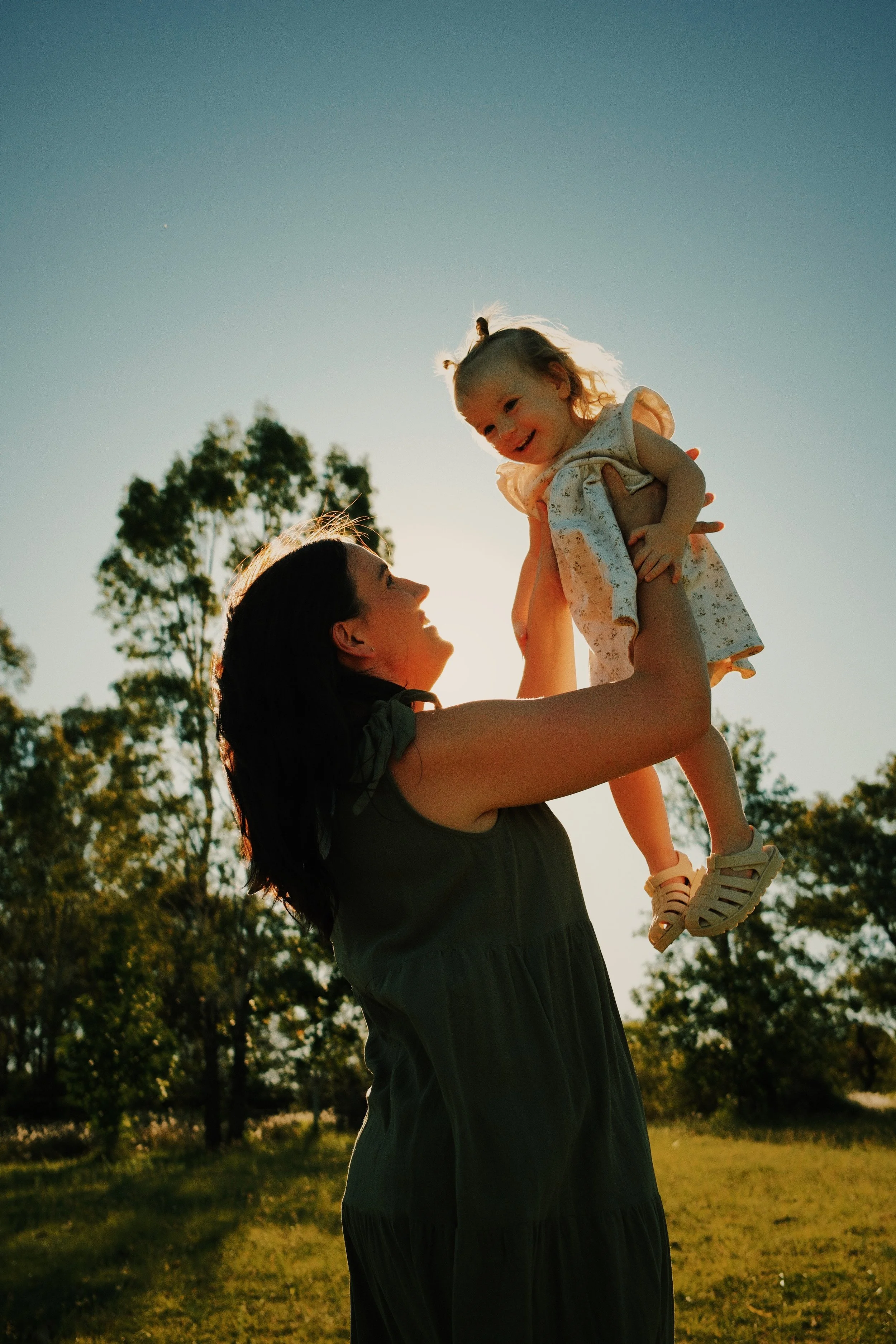 A woman lifting a young girl into the air outdoors during sunset, with trees and a clear sky in the background.