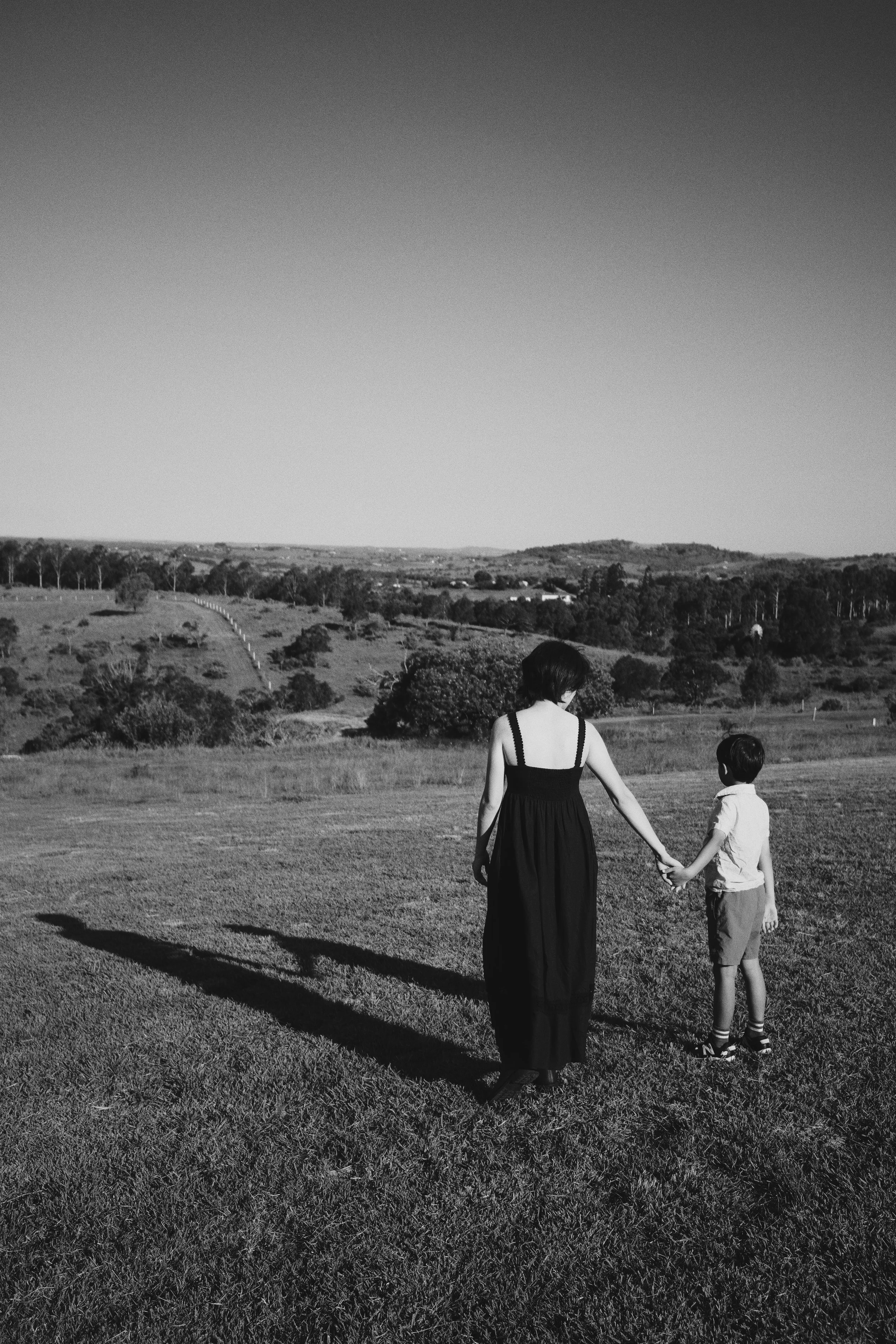 A woman and a boy holding hands and walking on a grassy field, with rolling hills and trees in the background under a clear sky.