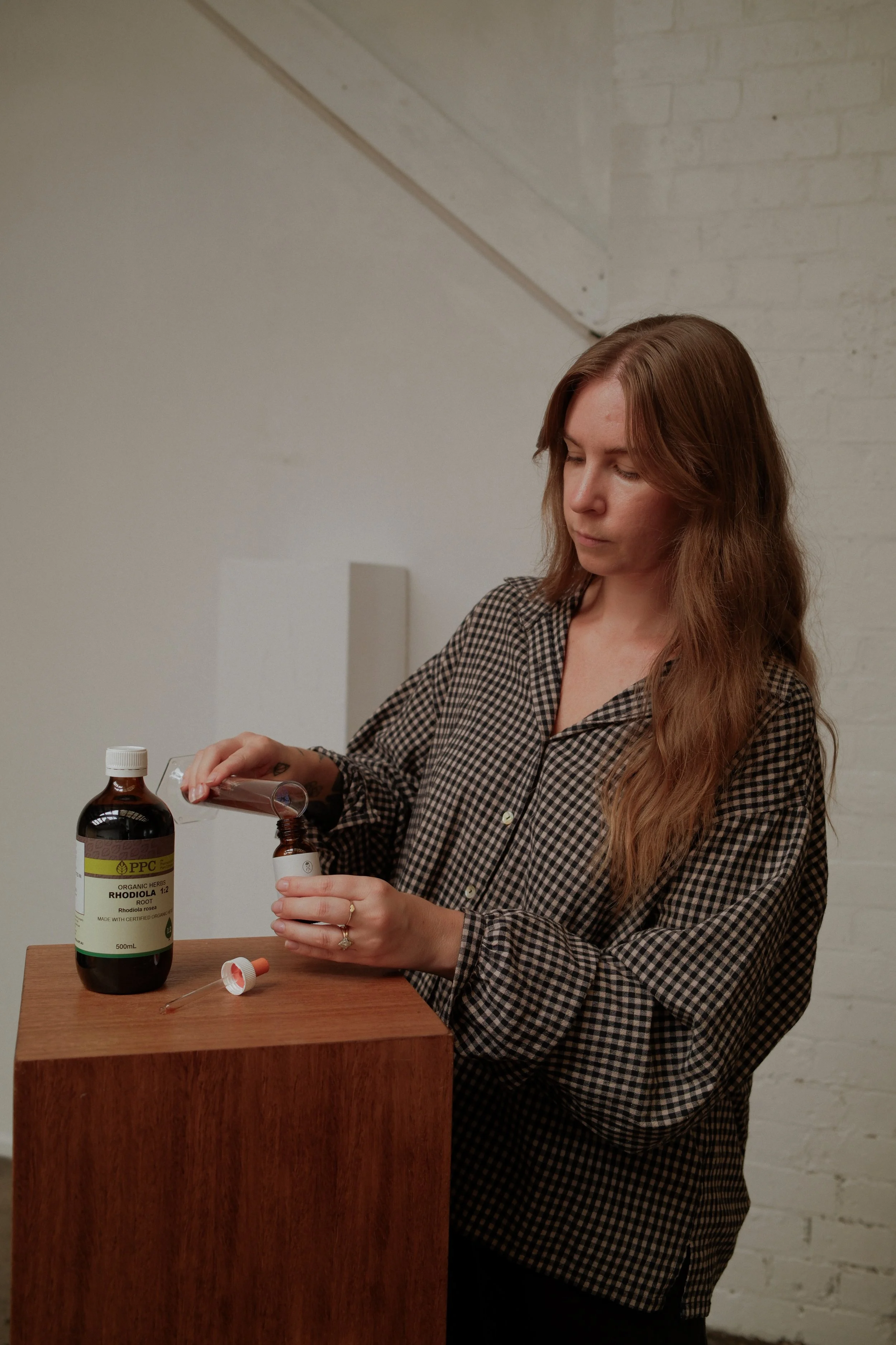 A woman with long, wavy reddish-brown hair wearing a black and white checkered shirt, standing at a wooden table, pouring liquid from a small dropper bottle into a small brown glass container.