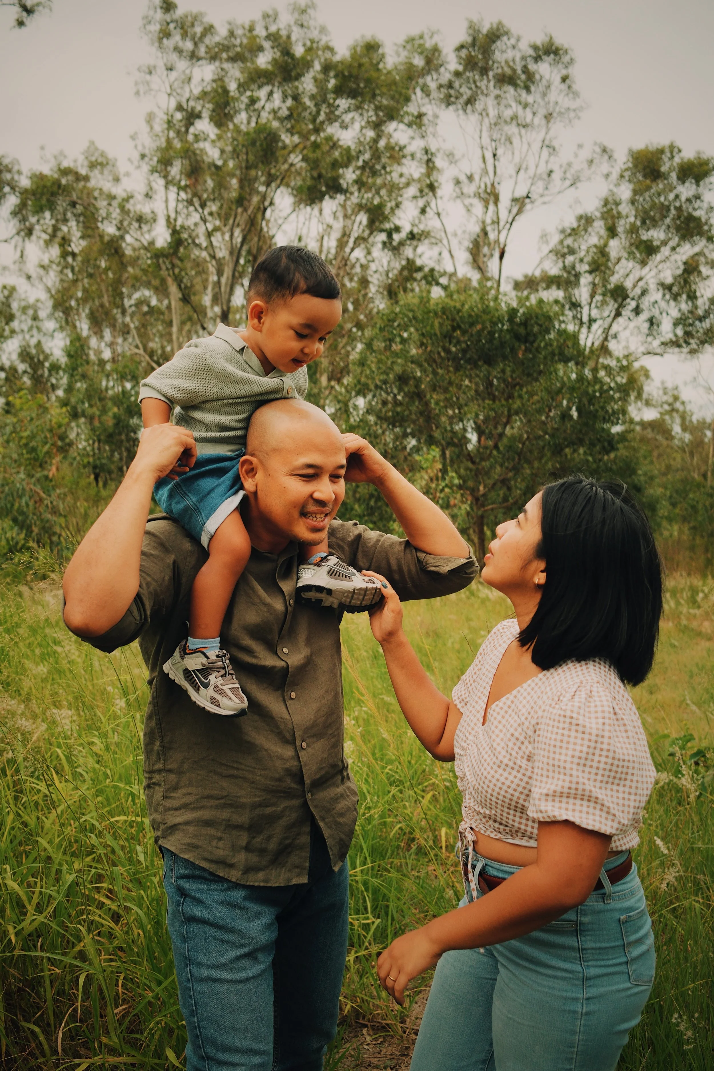 A family outdoors in a grassy field; a man carries a young boy on his shoulders, and a woman touches the man's face, all smiling and engaged.