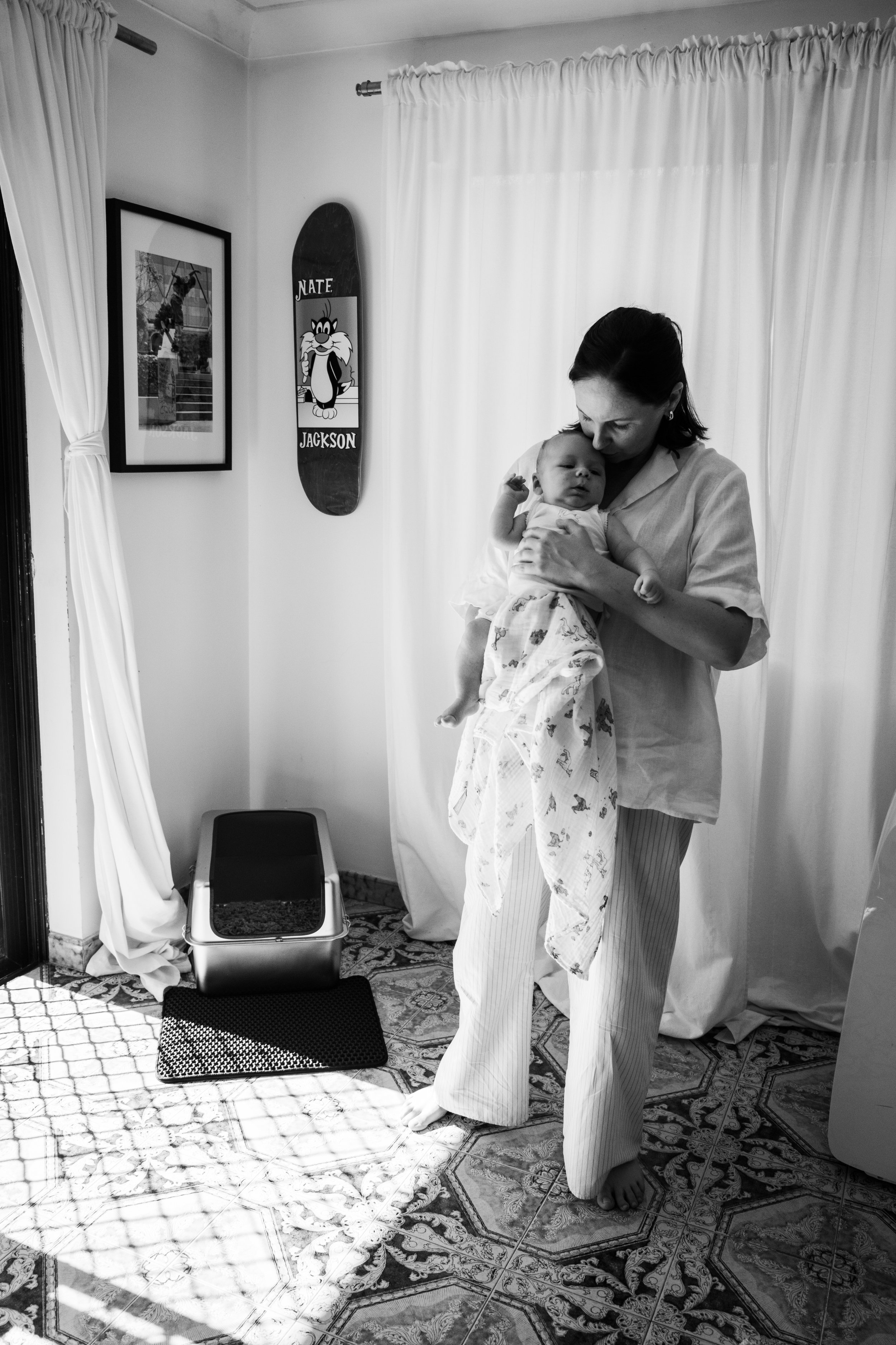 A woman holding a baby indoors with curtains behind and a skateboard with cartoon character on the wall.