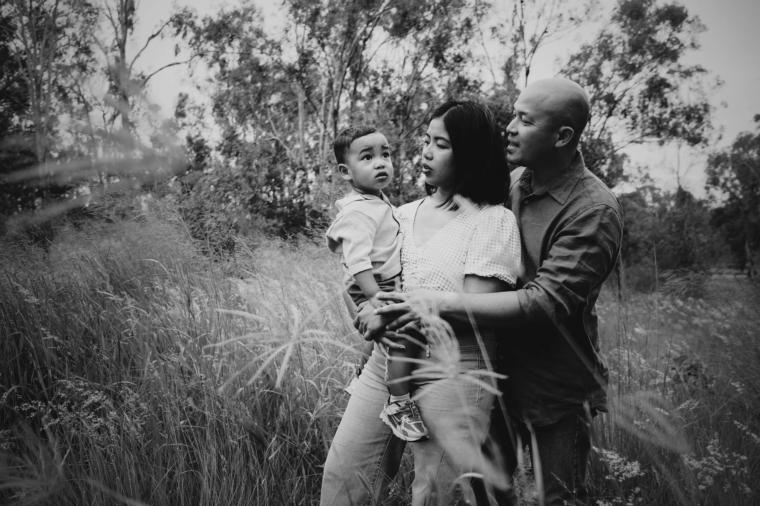 A family of three standing in tall grass outdoors, with trees in the background. The woman is holding a young boy, and the man stands beside them, all engaged in an intimate moment.