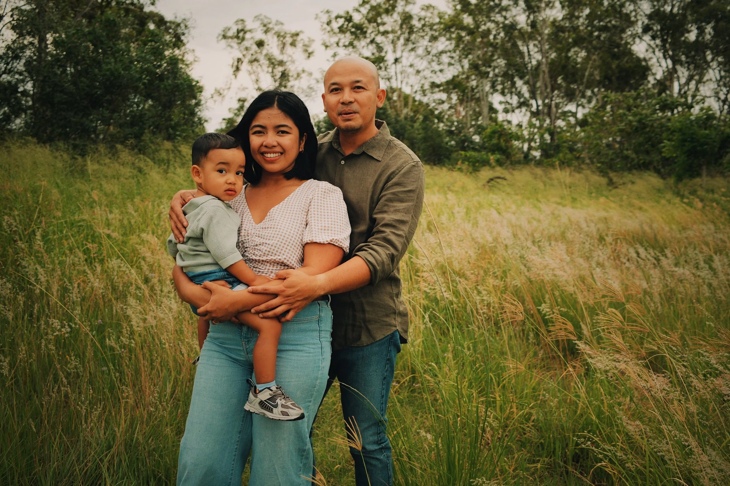 A family of three standing in a grassy field with trees in the background, smiling at the camera.