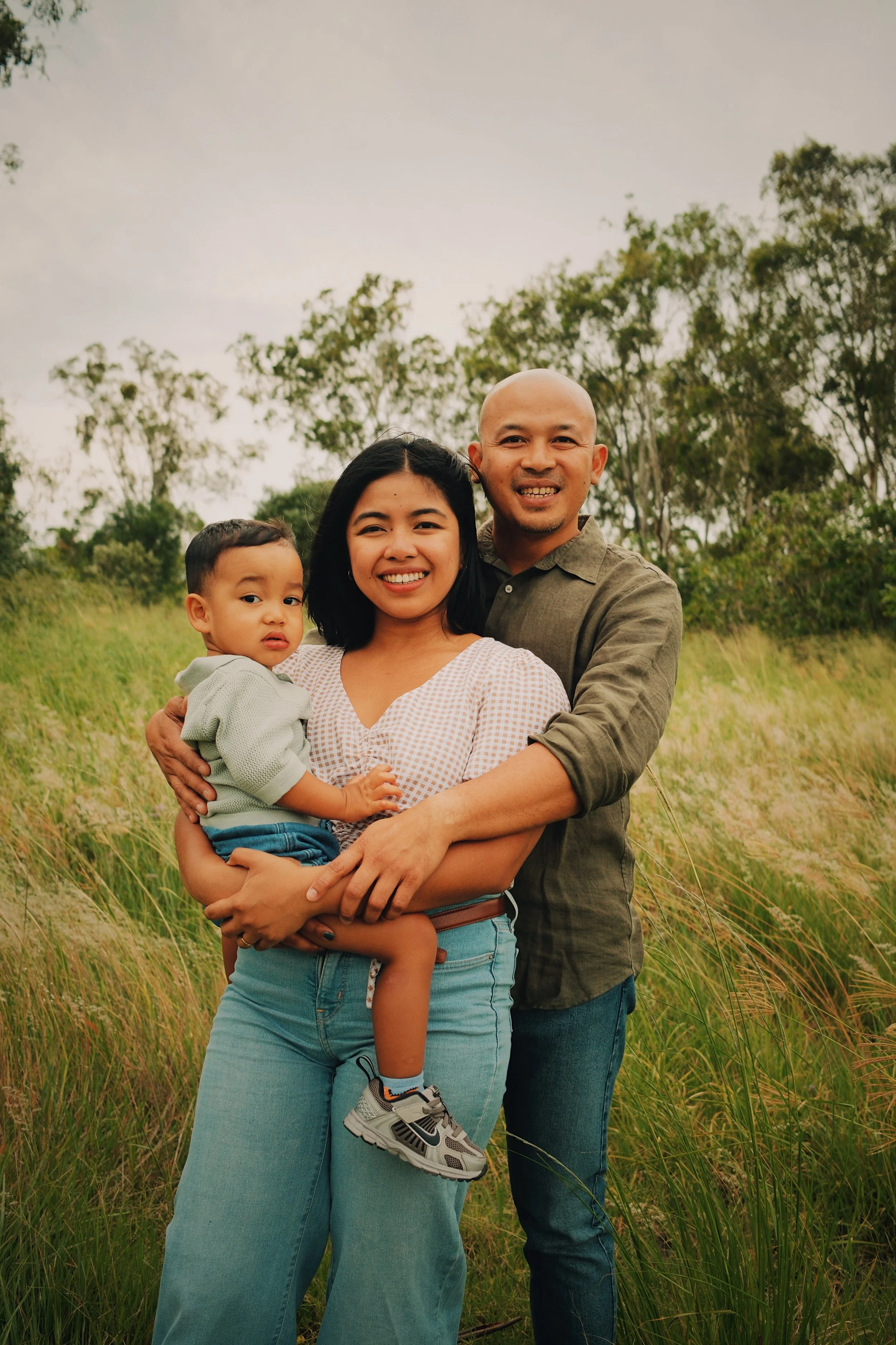 A smiling family of three standing outdoors in a grassy field with trees in the background.