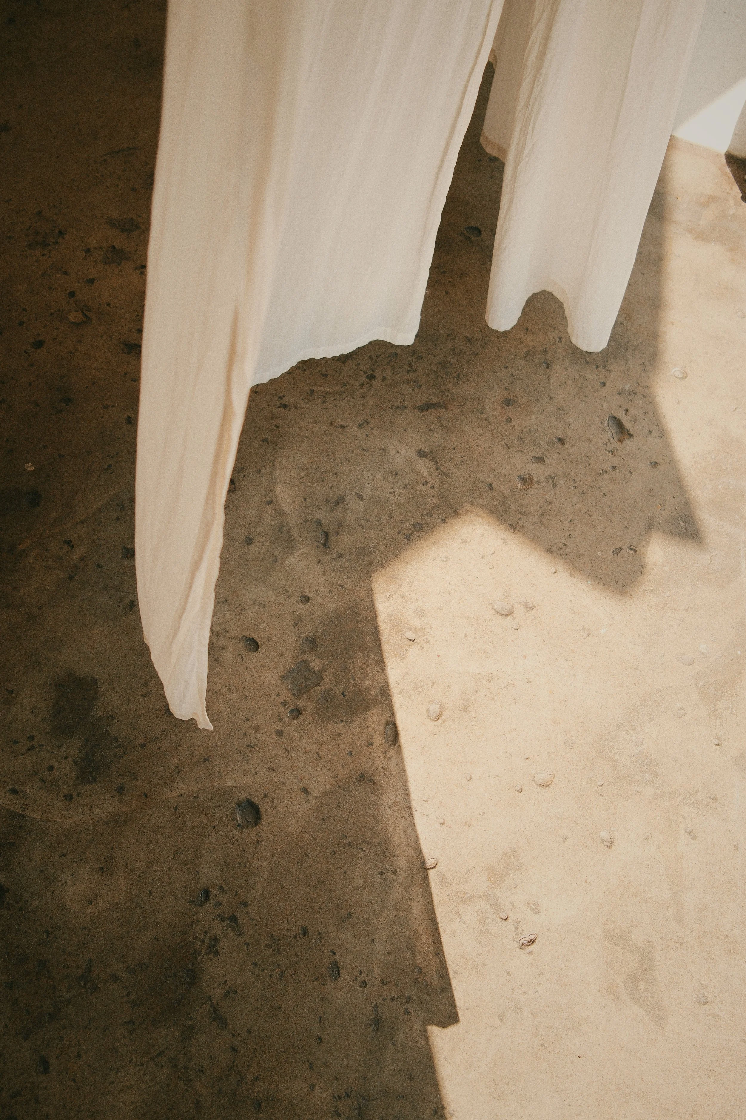 Close-up of a cream-colored curtain hanging above a concrete floor, with sunlight casting a shadow of the curtain onto the floor.