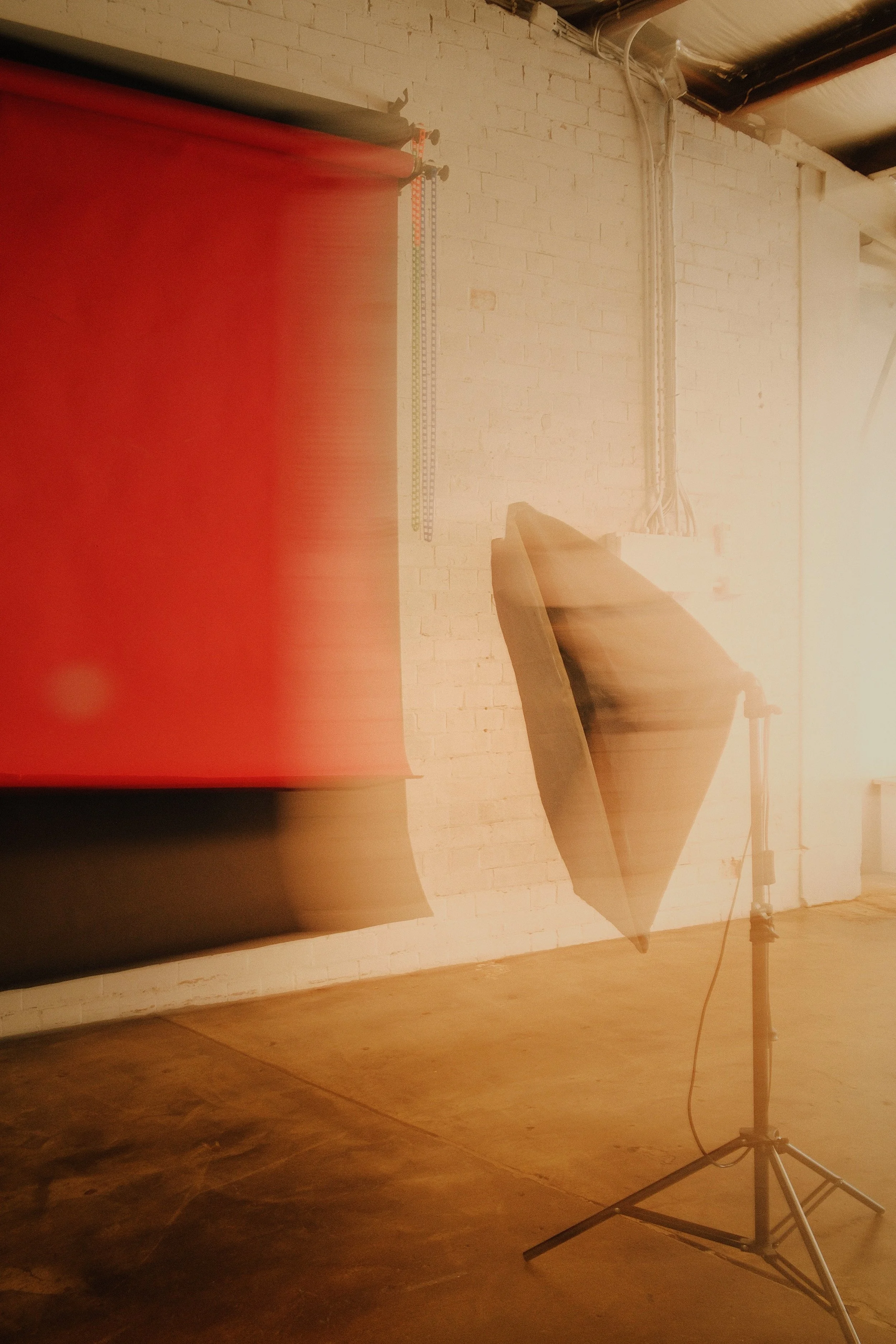 Studio setting with a brick wall background, a red backdrop on the left, and a large softbox studio light on a stand illuminating the scene.