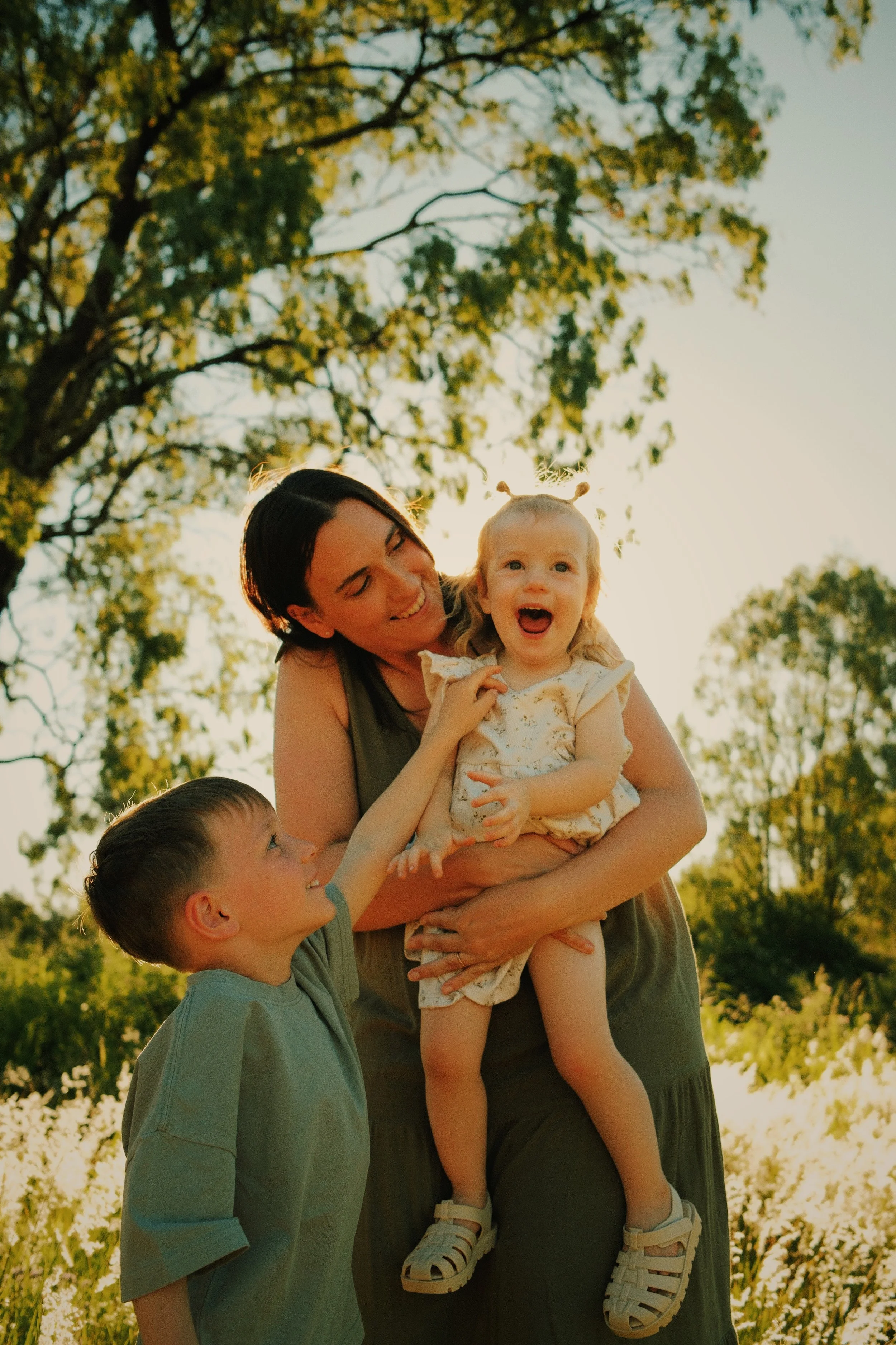 A smiling woman holding a young girl with pigtails, while a boy reaches towards her; they are outdoors in a field with trees and sunlight in the background.