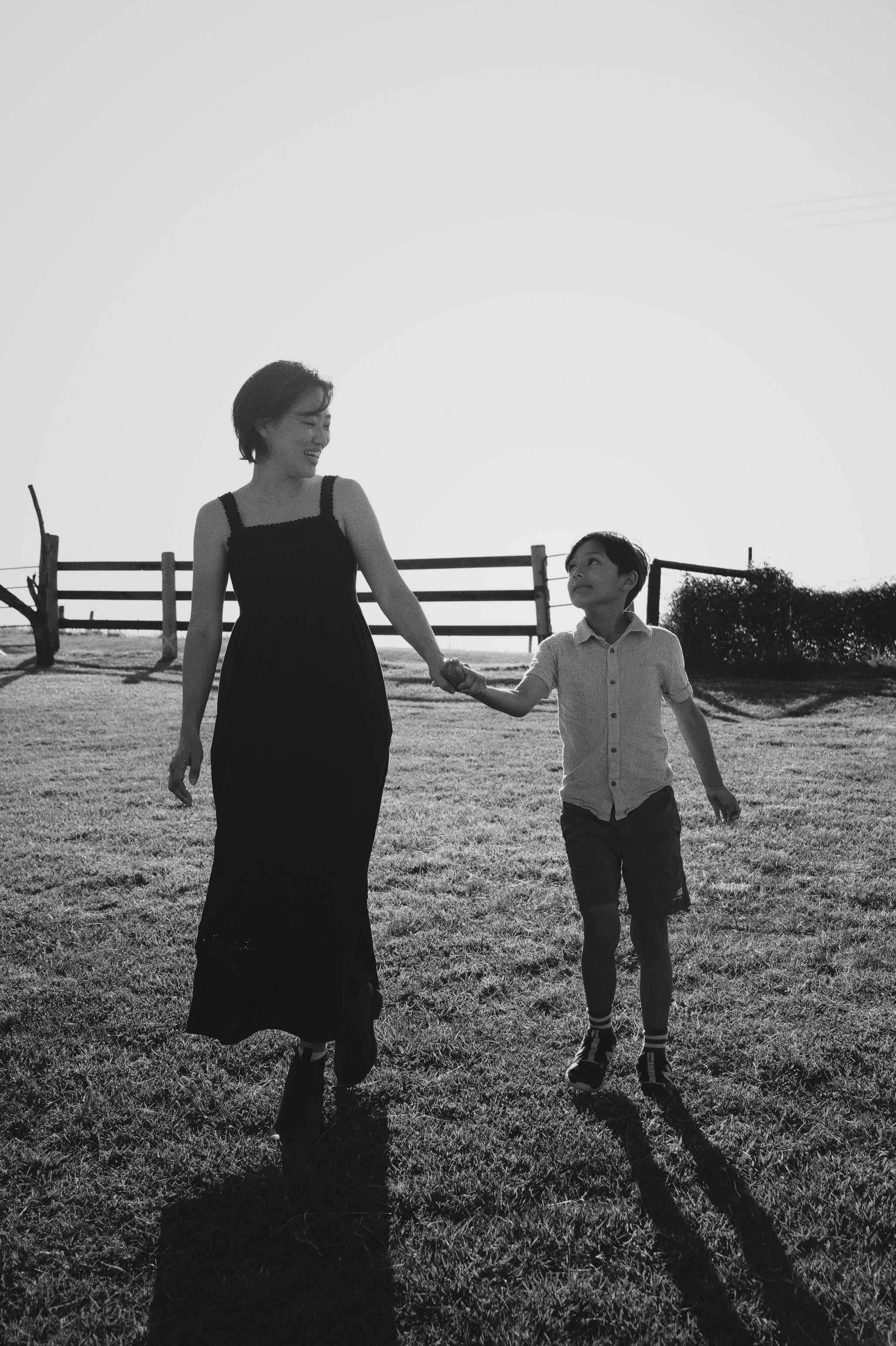 A woman and a boy holding hands and walking outside on grass during sunset. The woman is wearing a long dress and the boy is wearing a short-sleeved shirt and shorts. There is a fence and some bushes in the background.