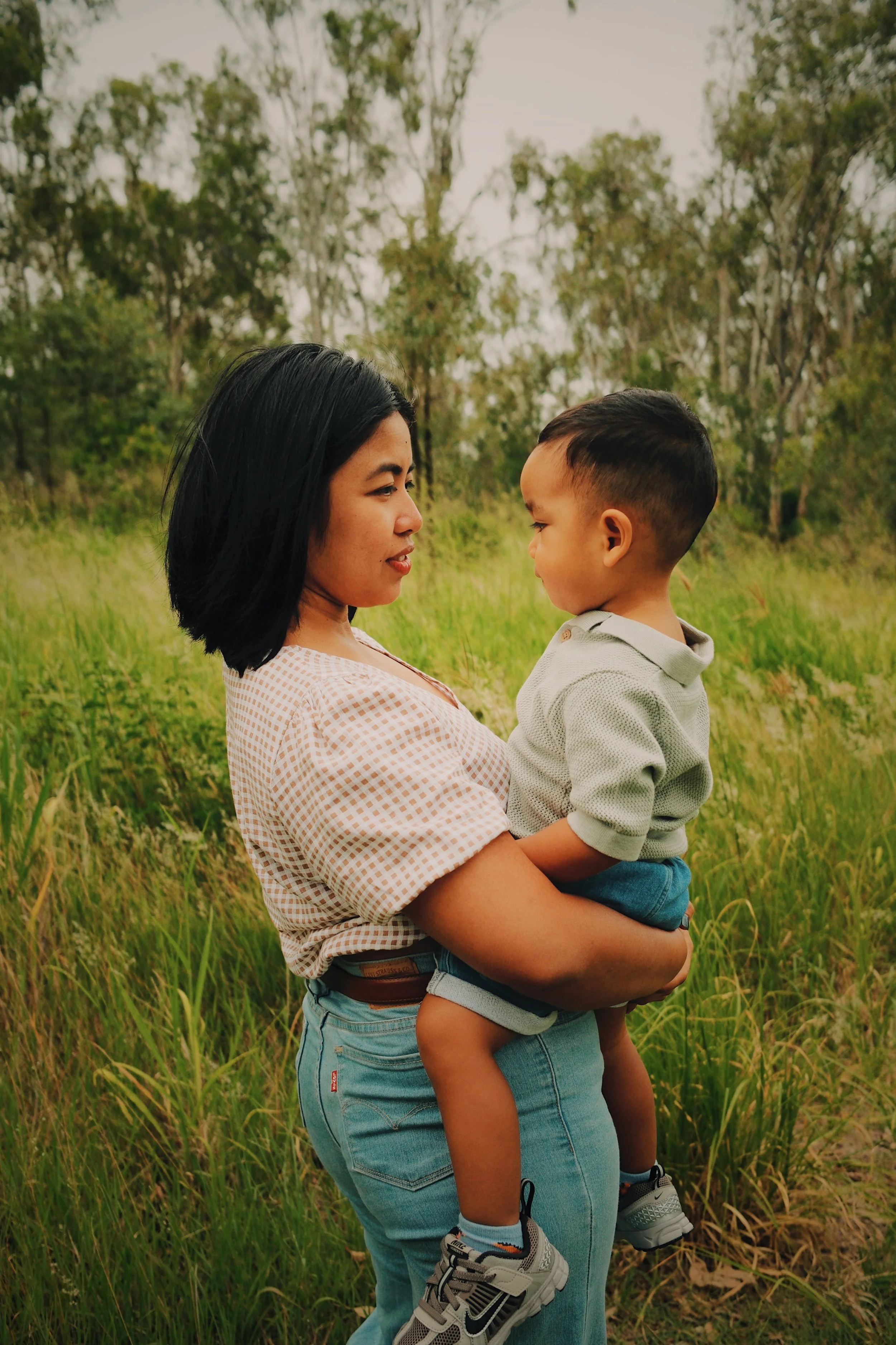 A woman holding a young boy in a green field with trees in the background, both facing each other