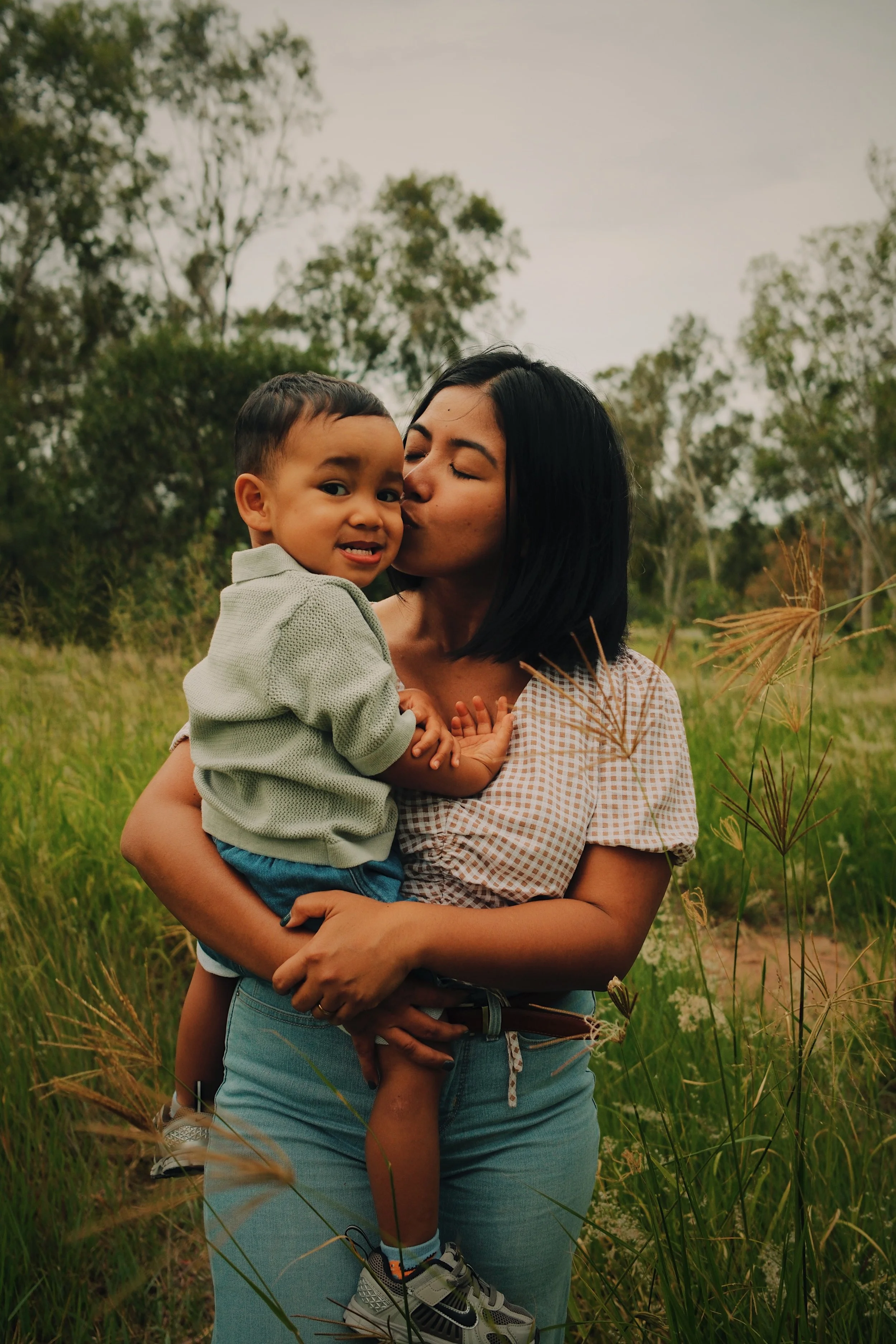 A woman holding a young boy in a grassy outdoor field, she kisses his cheek while he looks at the camera, surrounded by trees.