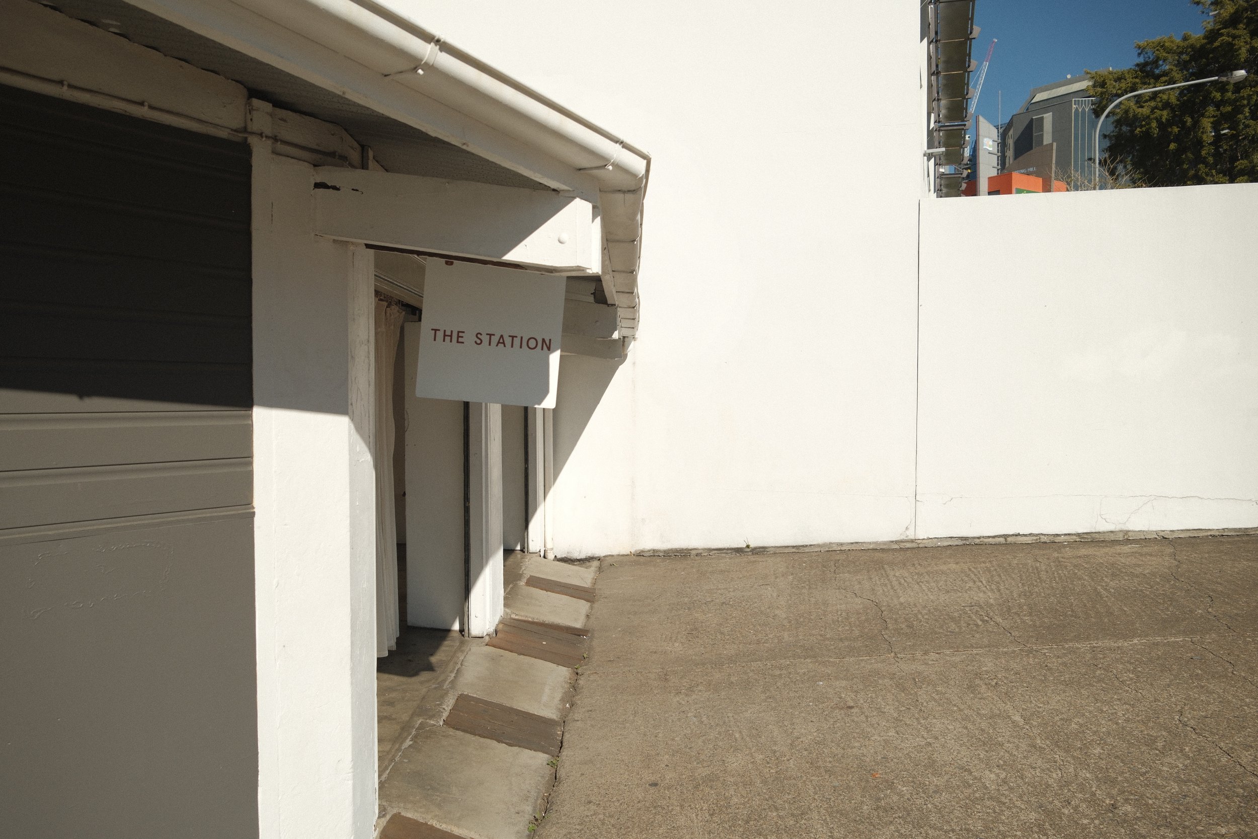 Entrance of a building with a sign that reads 'THE STATION' on a white wall, with a sidewalk and a large white wall in the background.