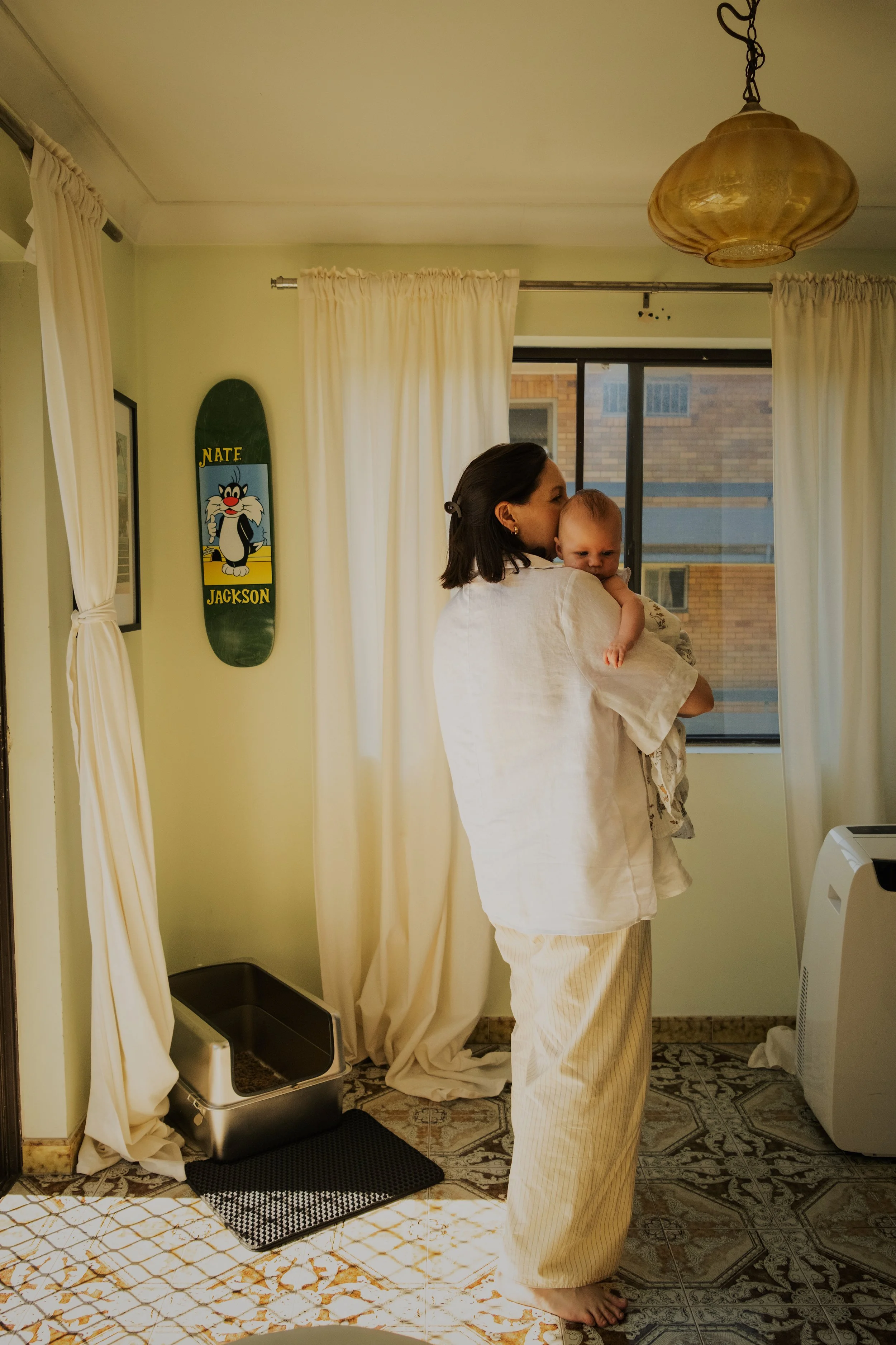 A woman holding a baby in a room with beige curtains and a window showing buildings outside. There is a skateboard with cartoon cat on the wall and a smart pet feeder on the floor.