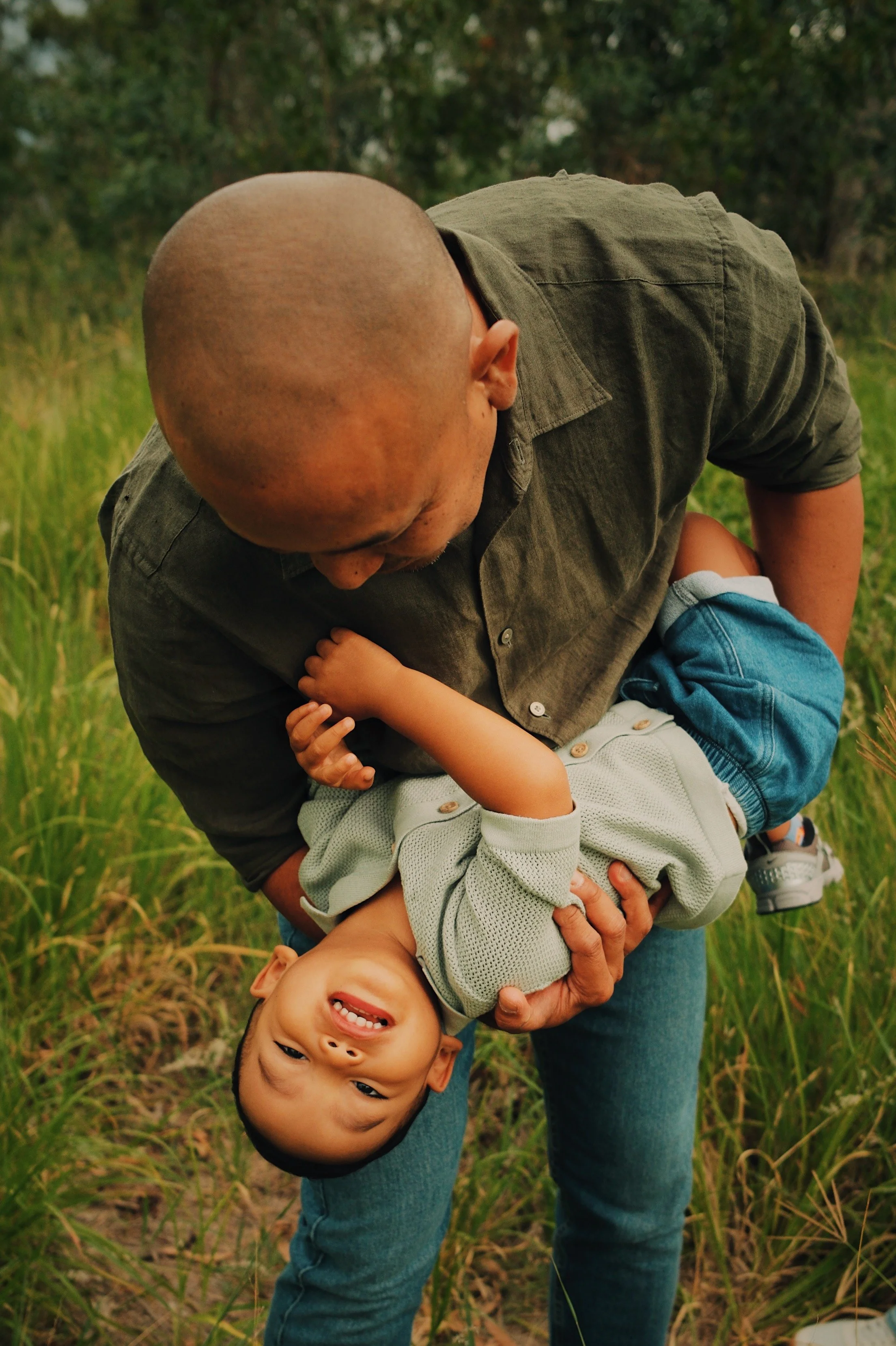 A man holding a smiling boy upside down in a grassy outdoor area.
