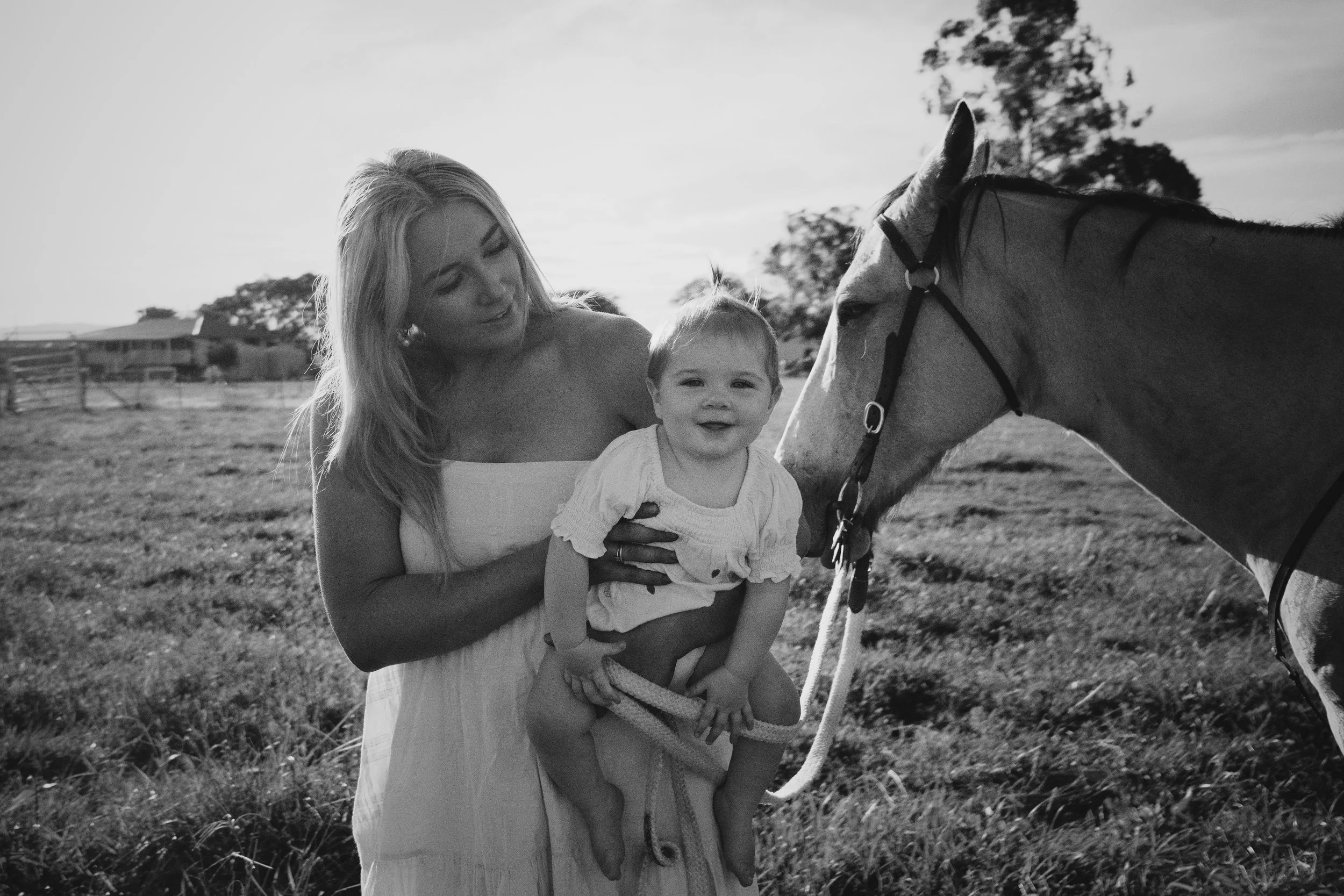 A woman holding a smiling toddler in front of a horse in an outdoor field, black and white photograph.