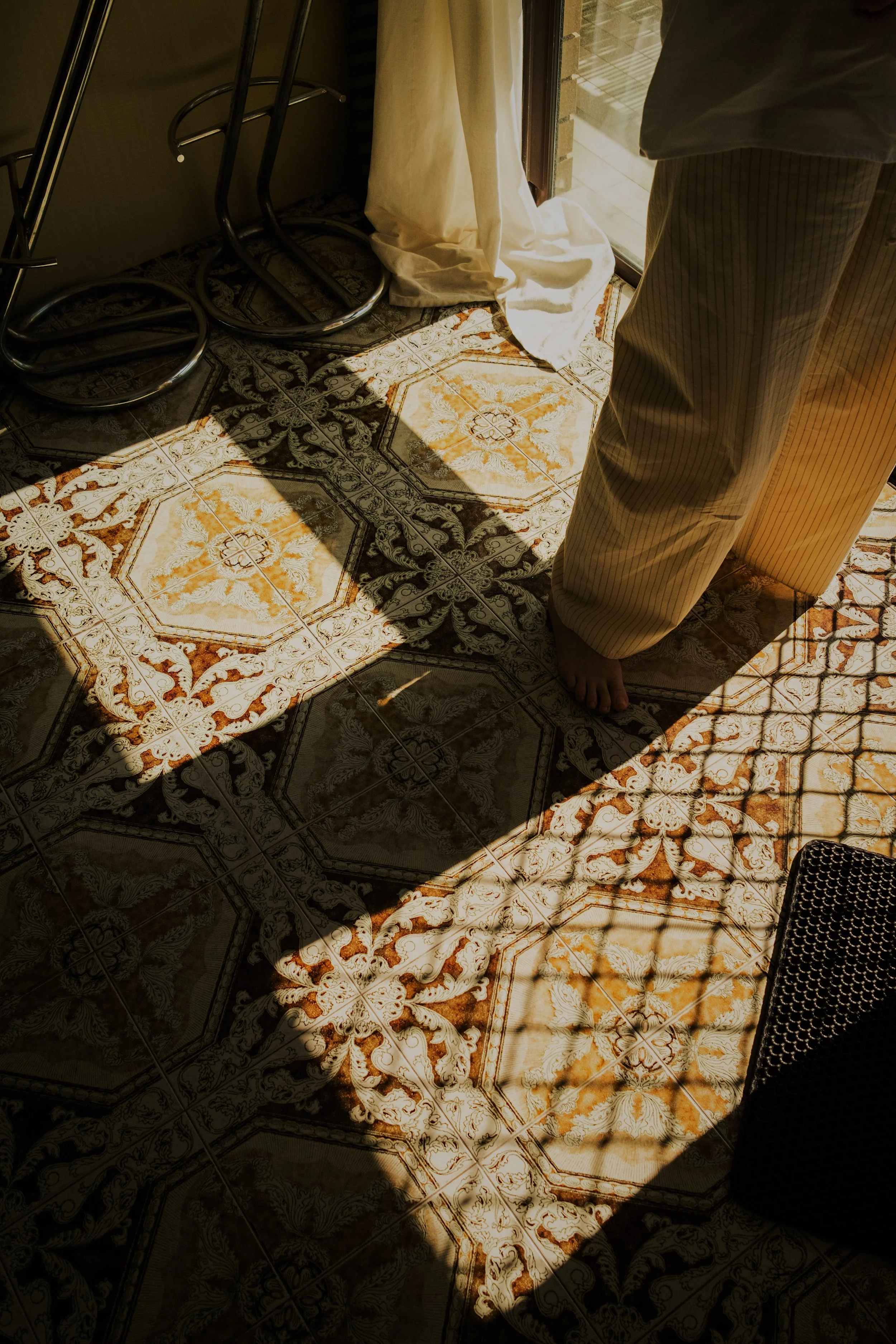 A person standing barefoot near a sliding glass door with patterned tile flooring and shadows cast by a metal window grille.