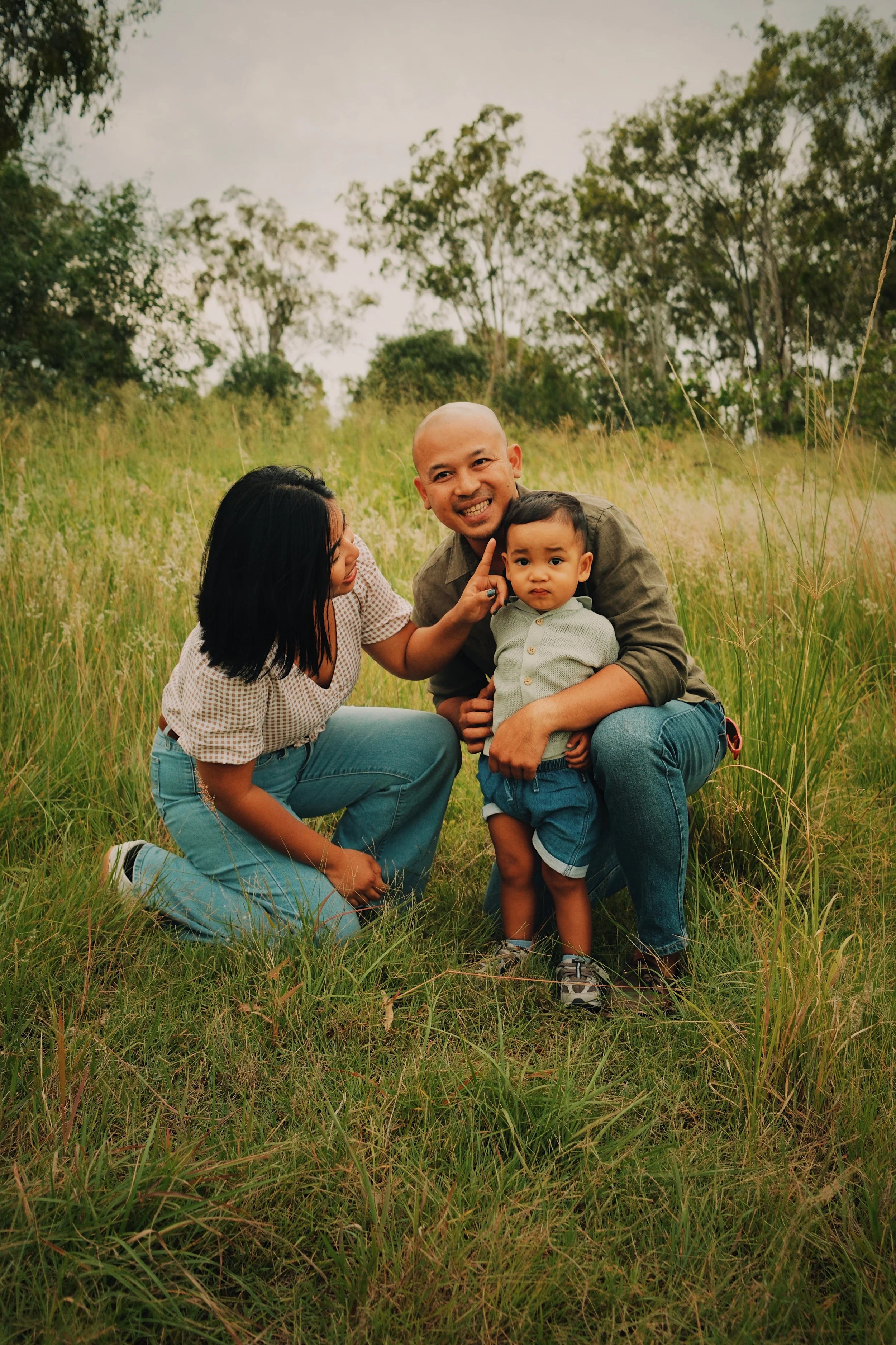 A family of three outdoors in a grassy field with trees in the background; a woman with black hair kneeling, a man smiling and holding a young boy, who has short dark hair and is wearing a light green shirt and blue shorts.