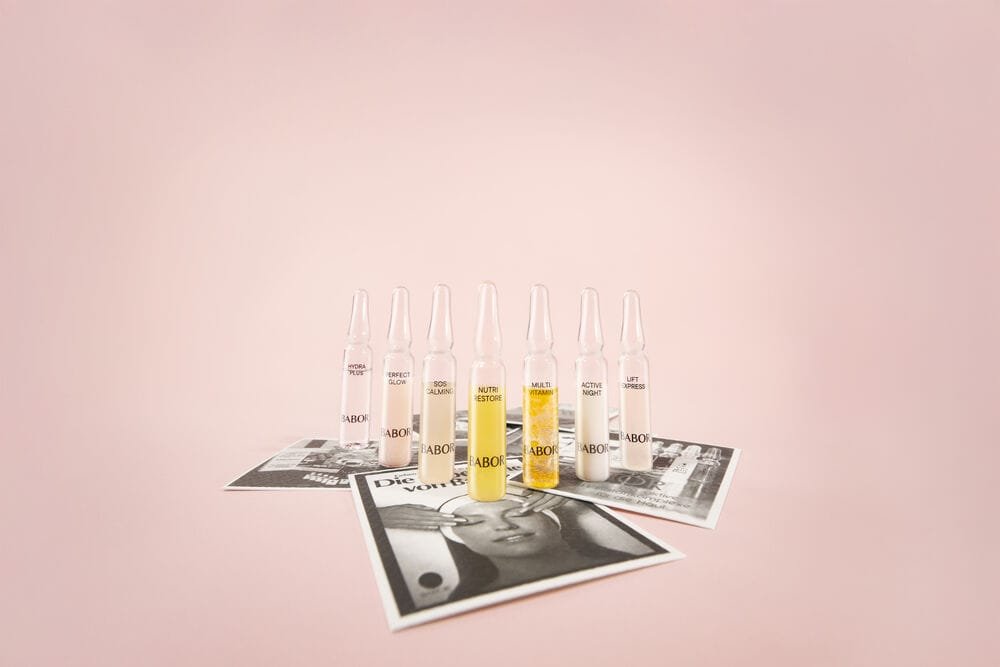 A row of 7 transparent bottles with liquid labeled 'BABOR', on top of posters with facial women care imagery, on a pink gradient background