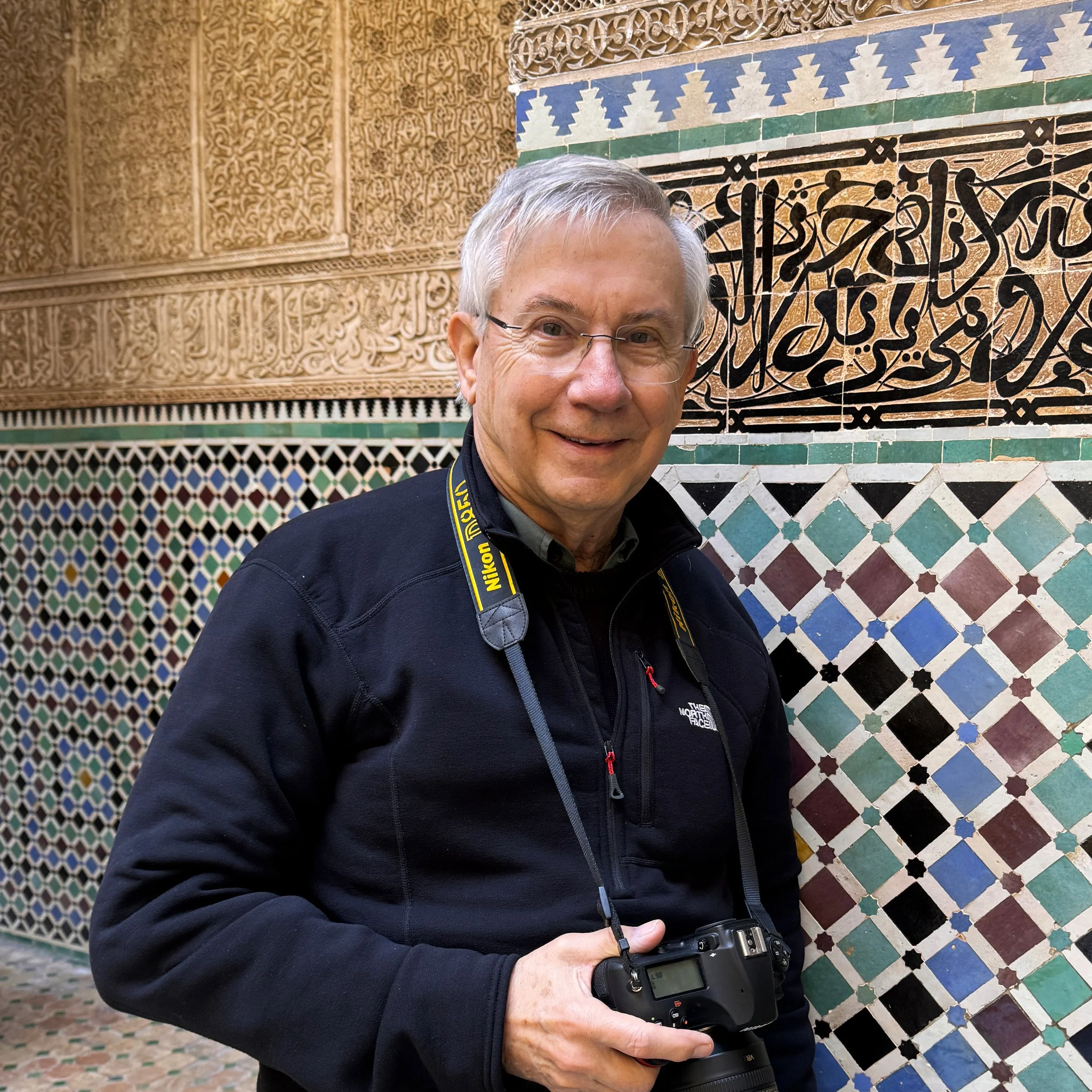 William Rawlings holding a camera and standing in front of a tan mosaic wall.