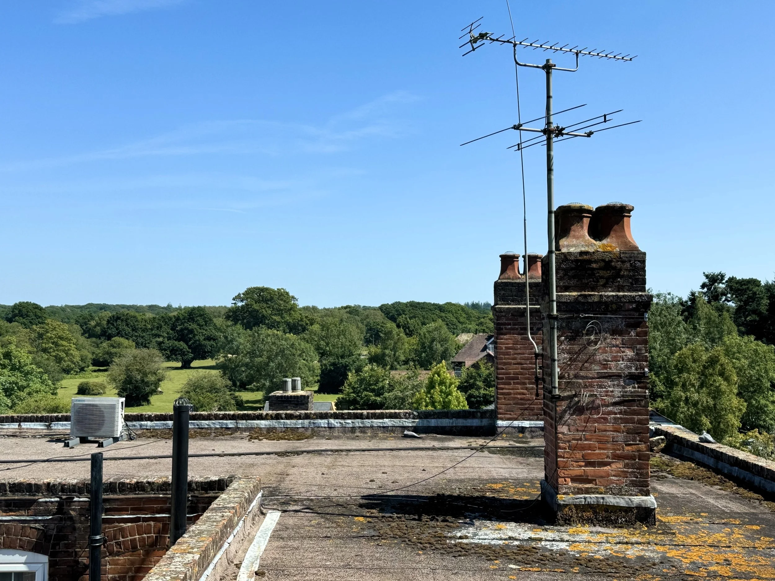View of a rooftop with a chimney, antennas, and an air conditioning unit, overlooking a green landscape with trees and a clear blue sky.