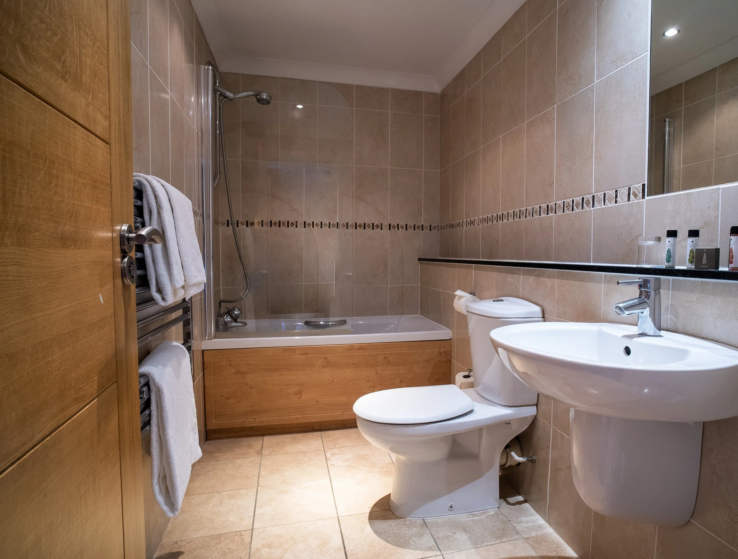 A bathroom with a toilet, a sink, and a bathtub with a shower, beige tiled walls and floor, and a wooden door.