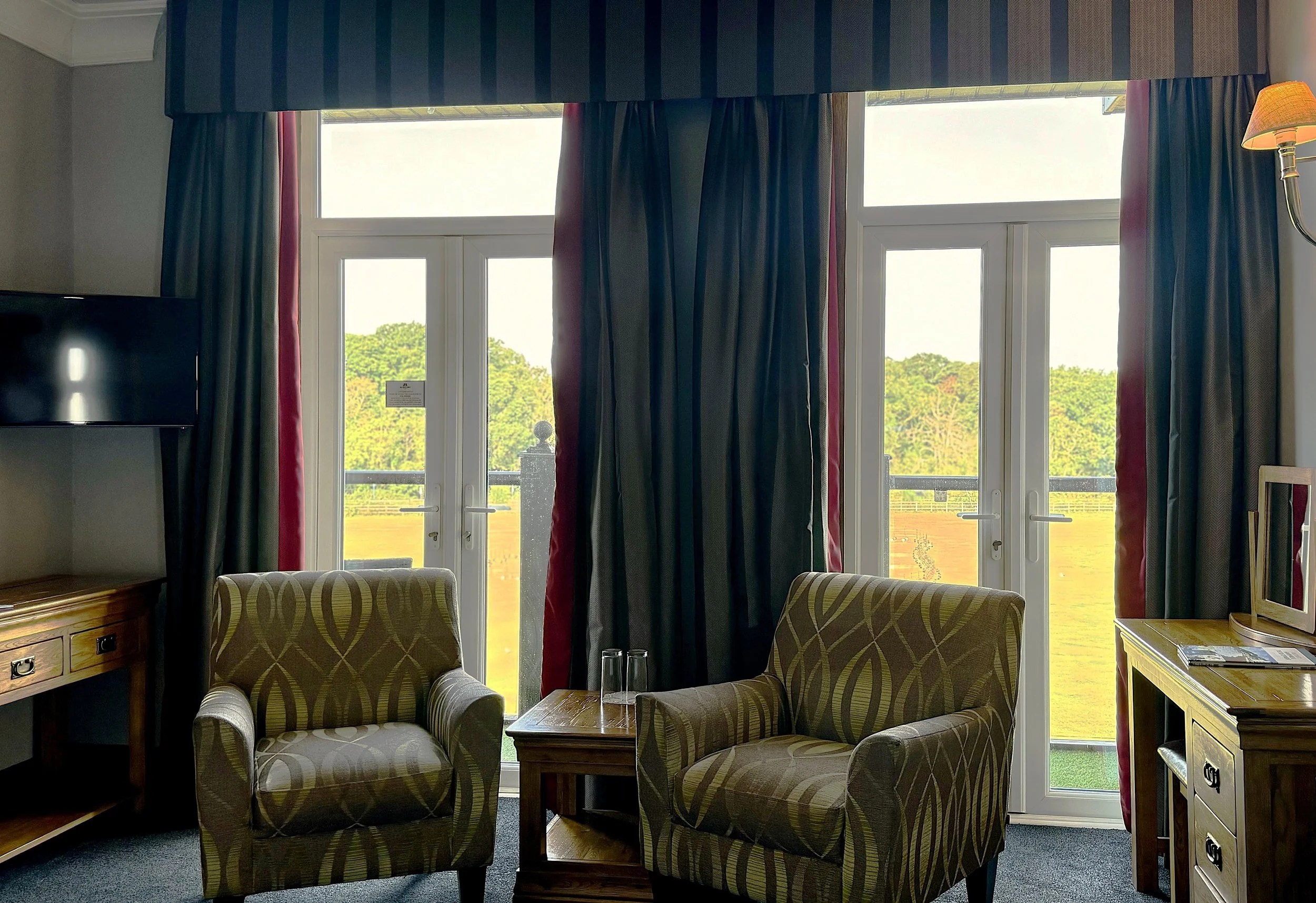 Interior of a room with two patterned armchairs, a wooden table with two glasses, a dark curtain, and large windows showing a green outdoor landscape.