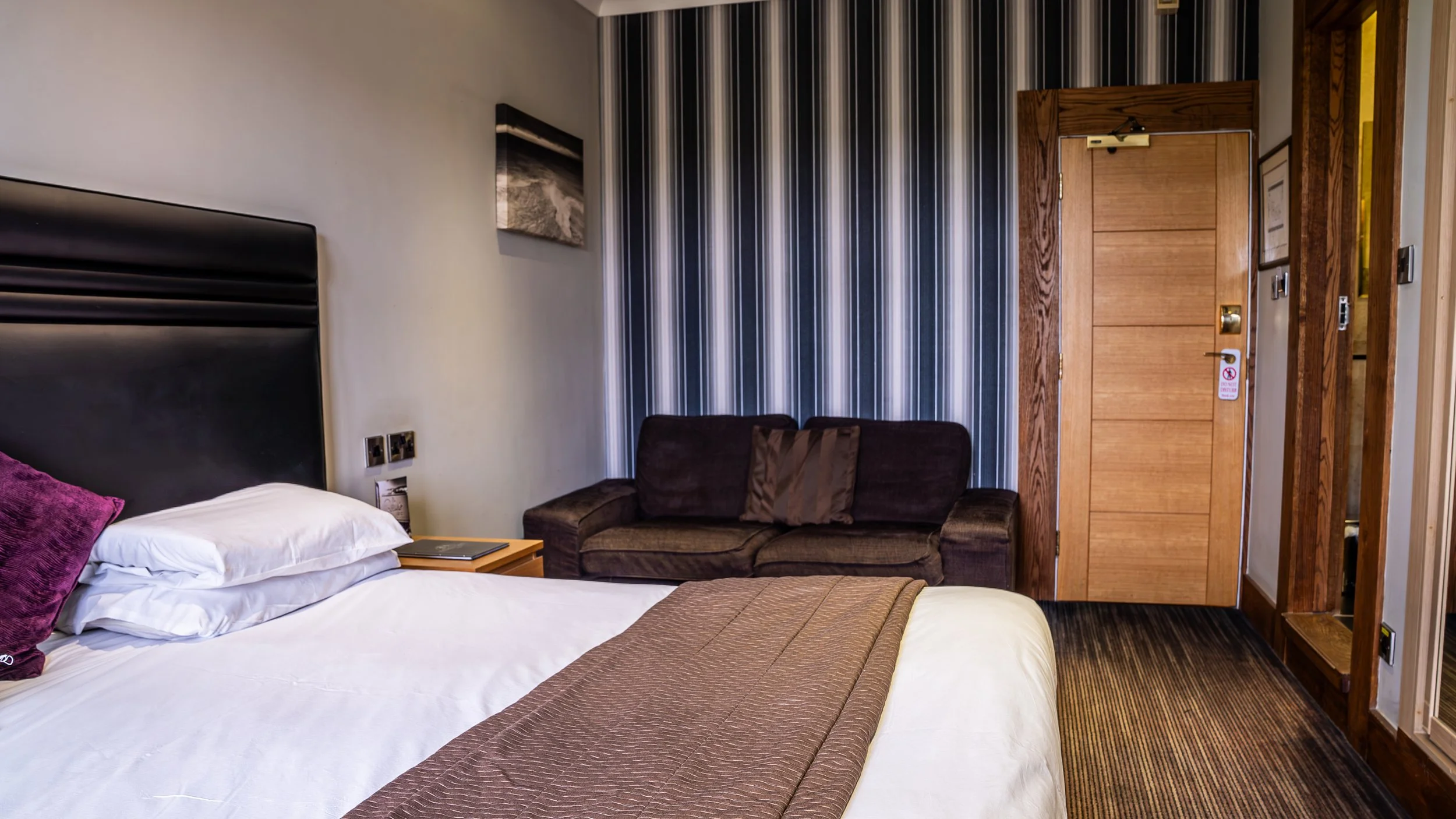 Hotel room with a bed, side table with book, a dark brown sofa with pillows, striped wallpaper, wooden door, and part of a mirror.