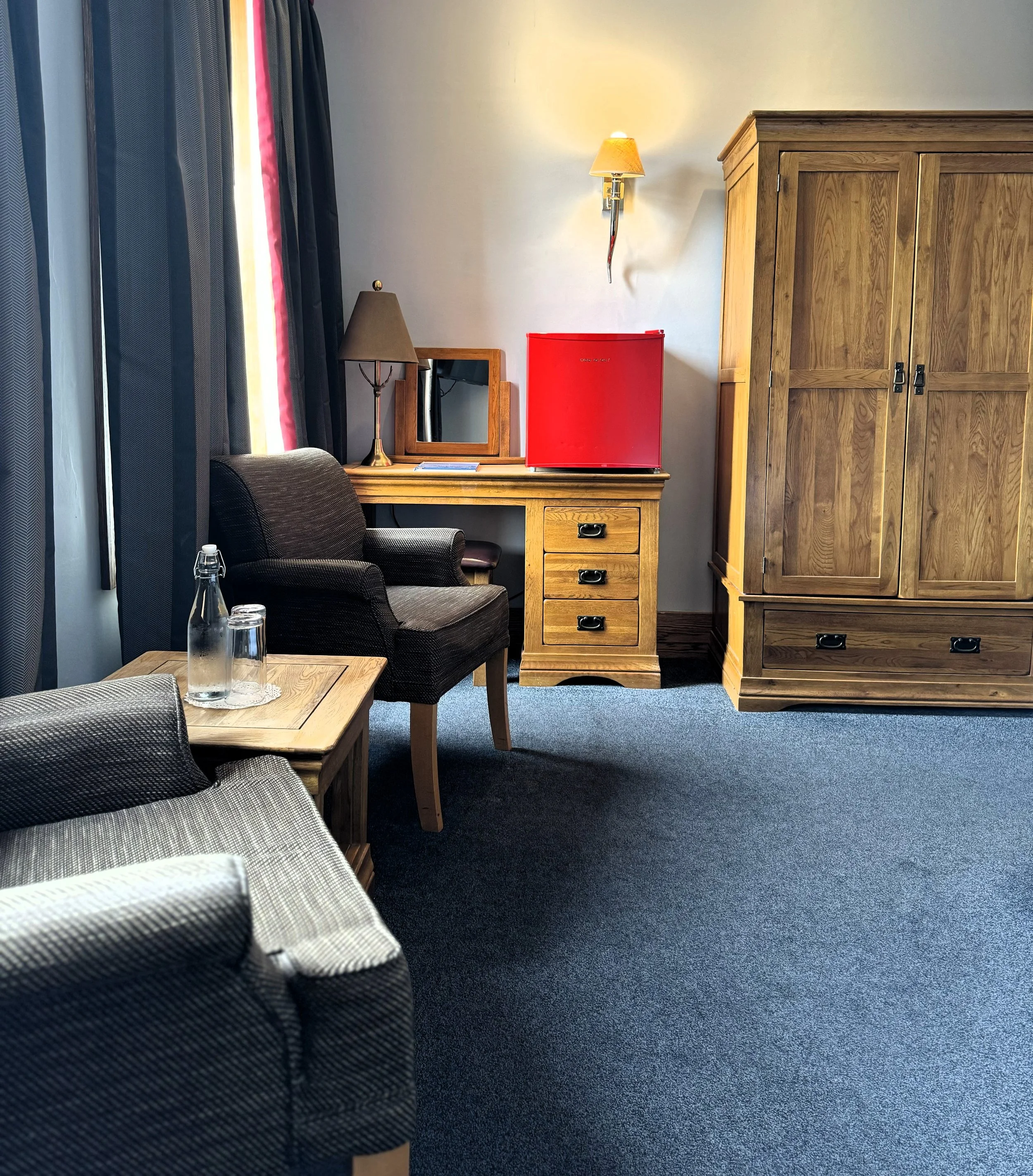 A room with a dark blue carpet, a wooden desk with a red mini fridge on top, a small mirror, and a table lamp. There are two dark upholstered chairs, a small wooden side table with a glass water bottle, and a large wooden wardrobe. There is a window 