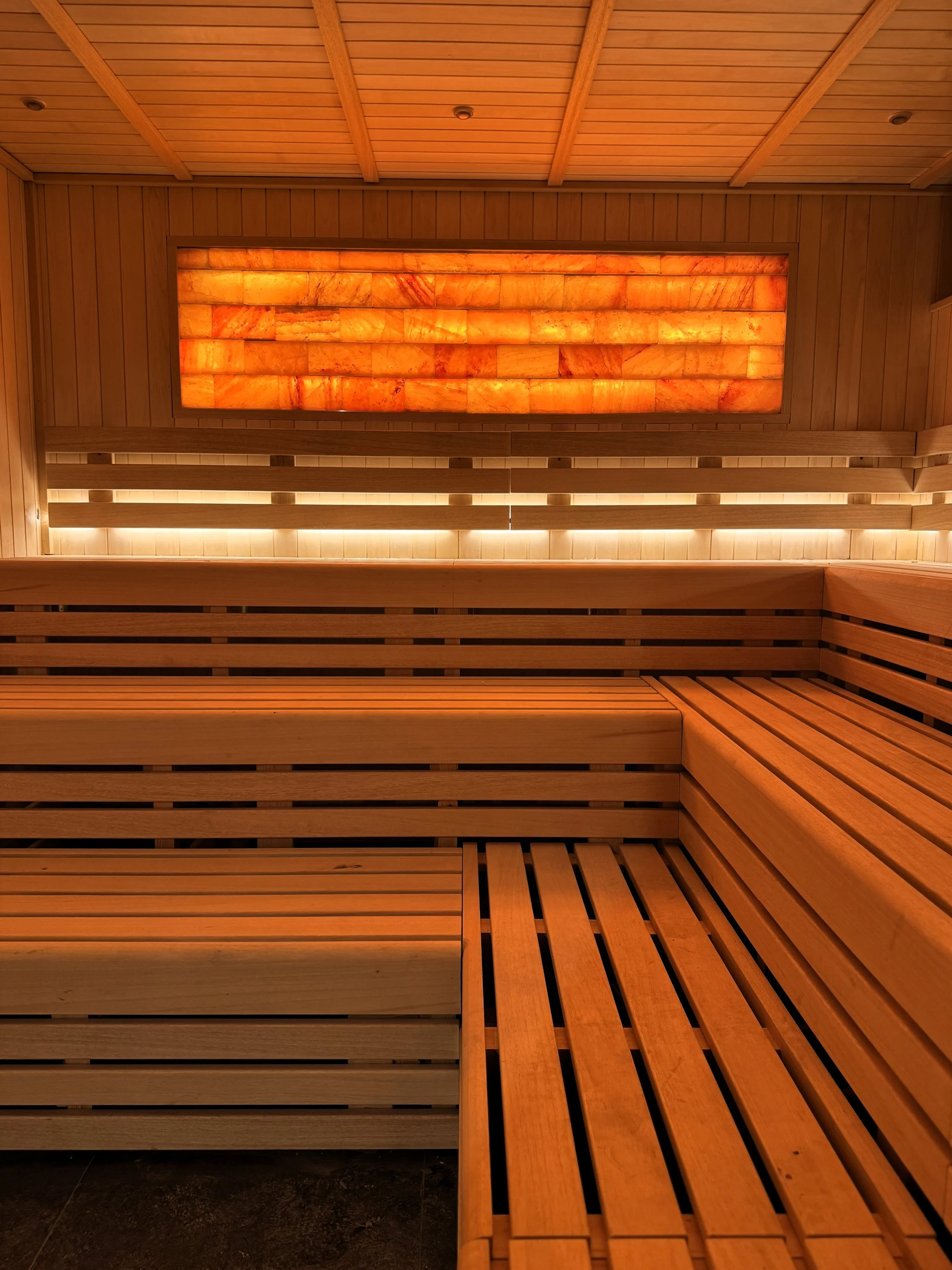 A warm, softly lit wooden sauna interior featuring smooth horizontal slatted benches and a glowing Himalayan salt brick panel mounted on the back wall.